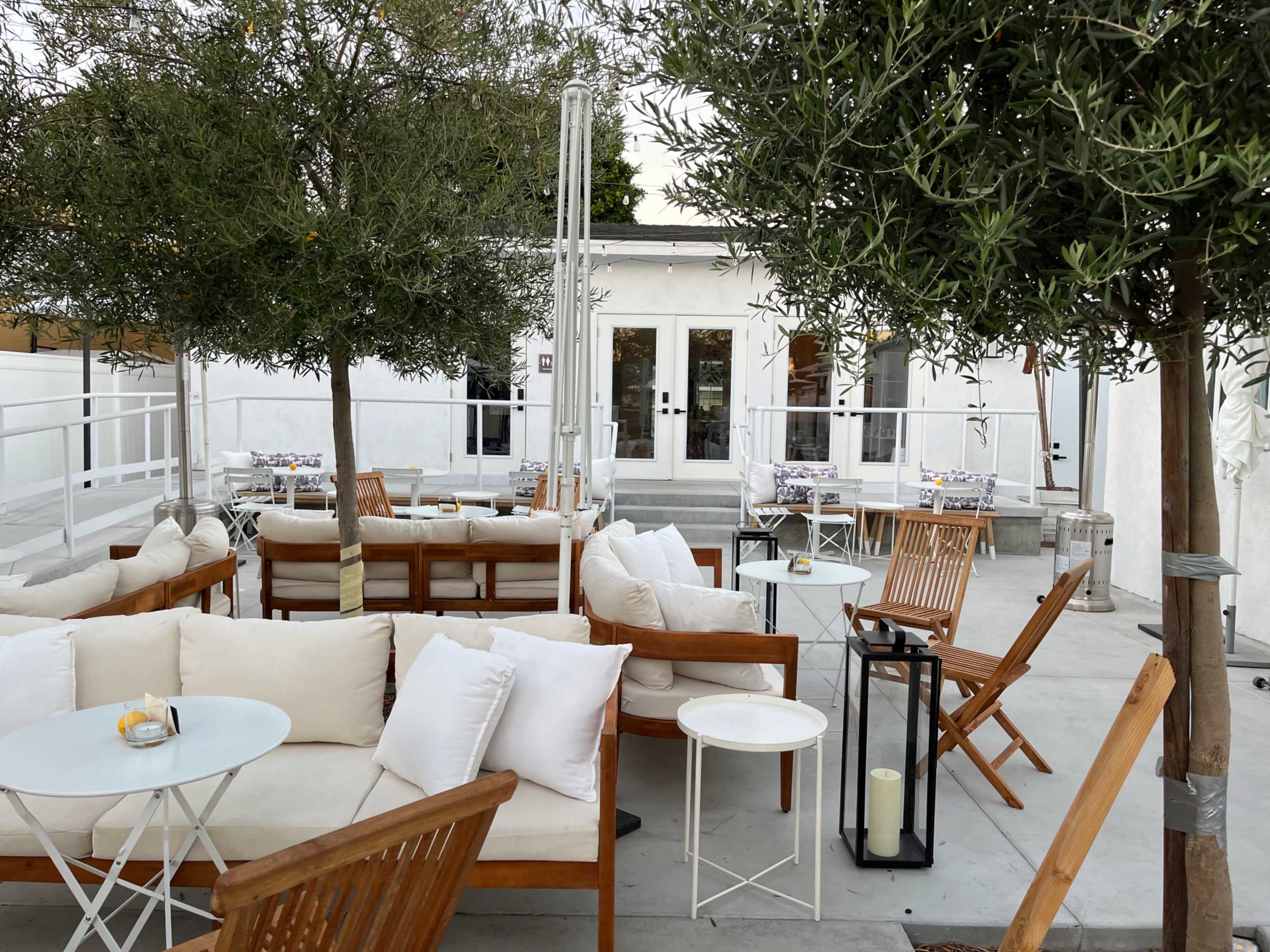 A modern outdoor seating area with wooden furniture, white cushions, and potted trees, set against a white-walled building.