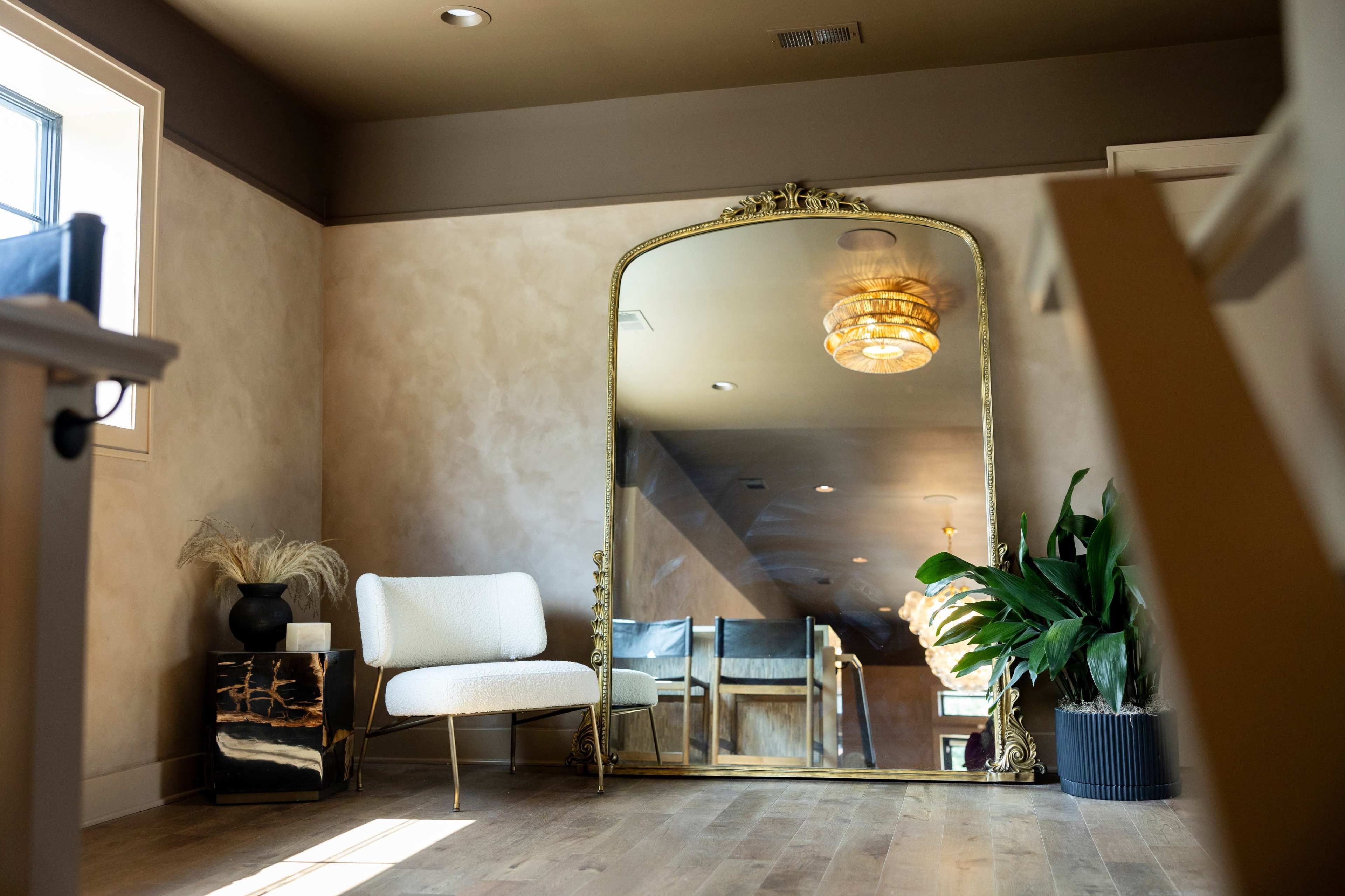 A spacious room with a large ornate mirror, a white upholstered chair, a black and gold side table, and a potted plant, all set against a softly textured wall.