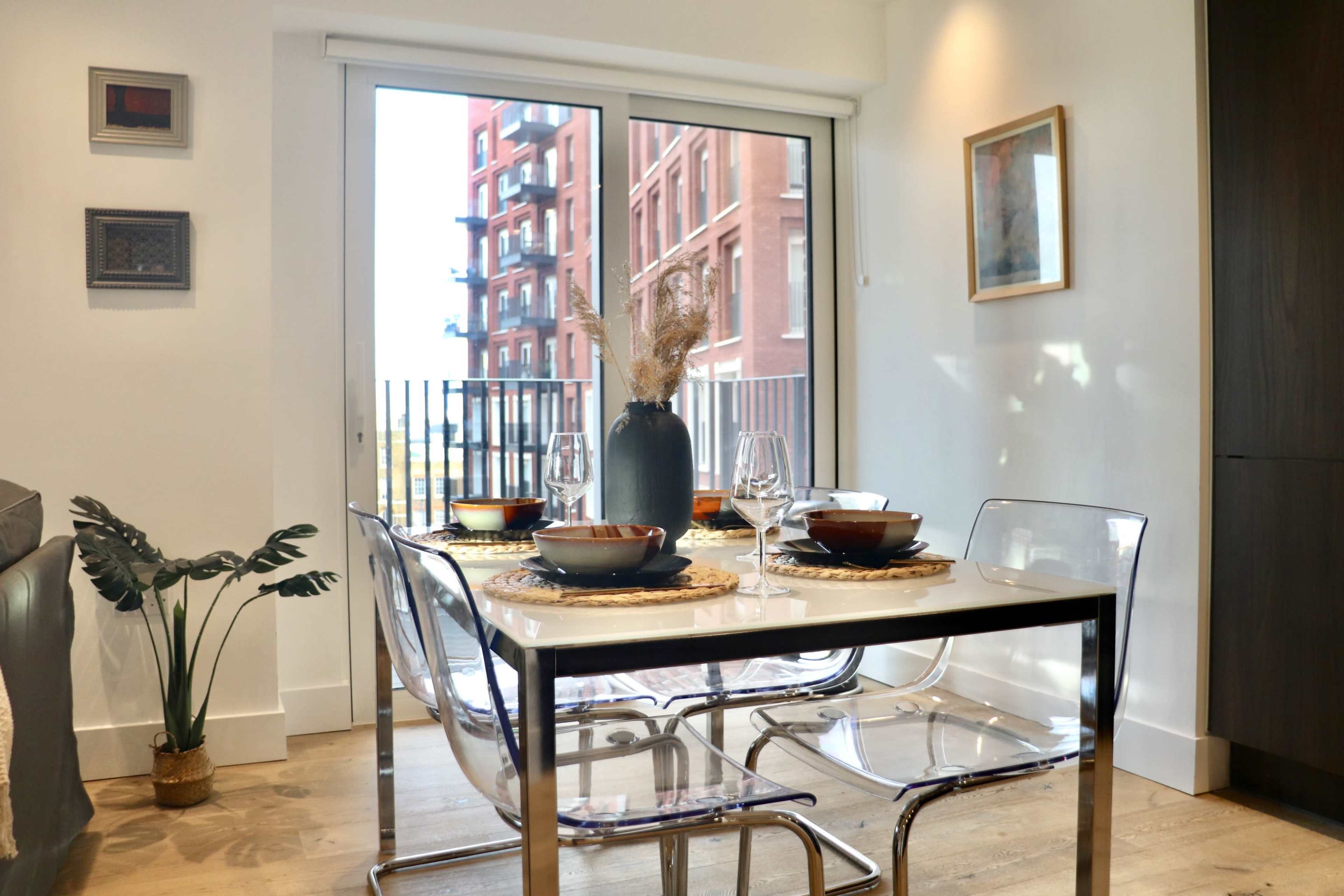 A dining table set for four is arranged next to a large window overlooking a building with red brick exterior.