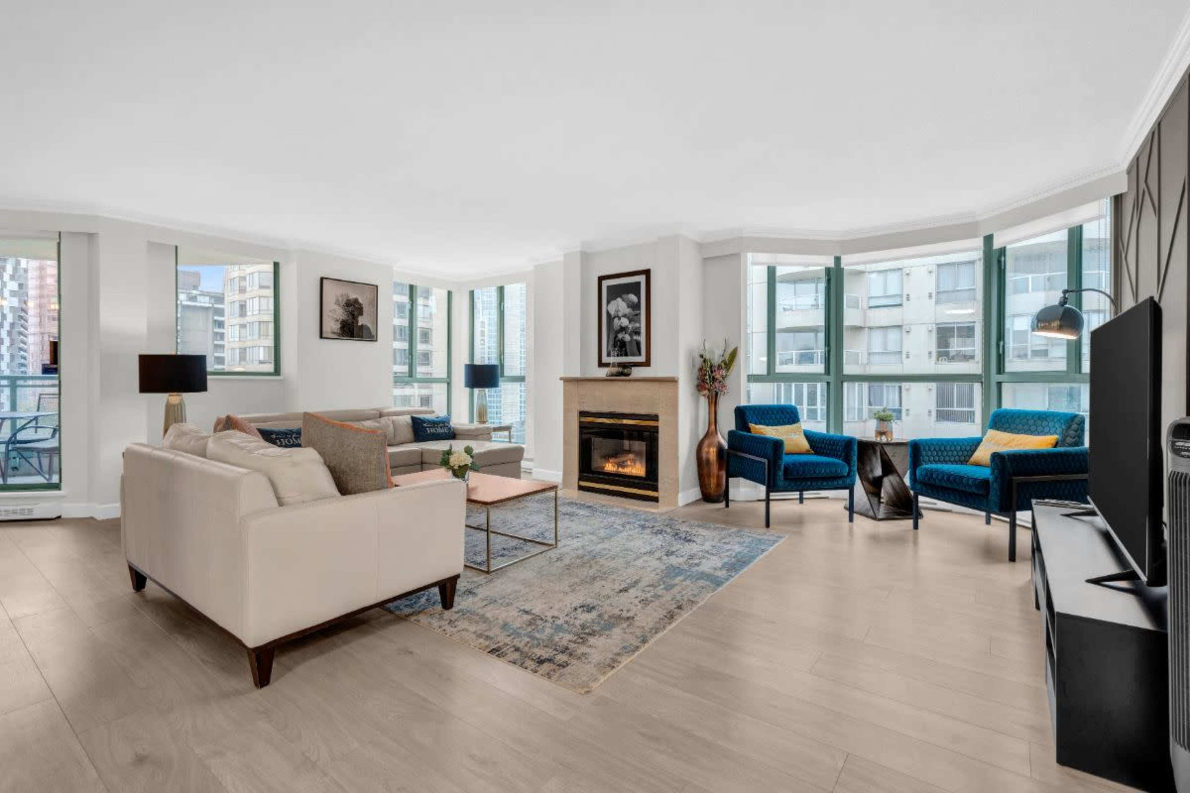 The image shows a modern living room with large windows, a fireplace, two blue chairs, a beige sofa, and light wooden flooring.