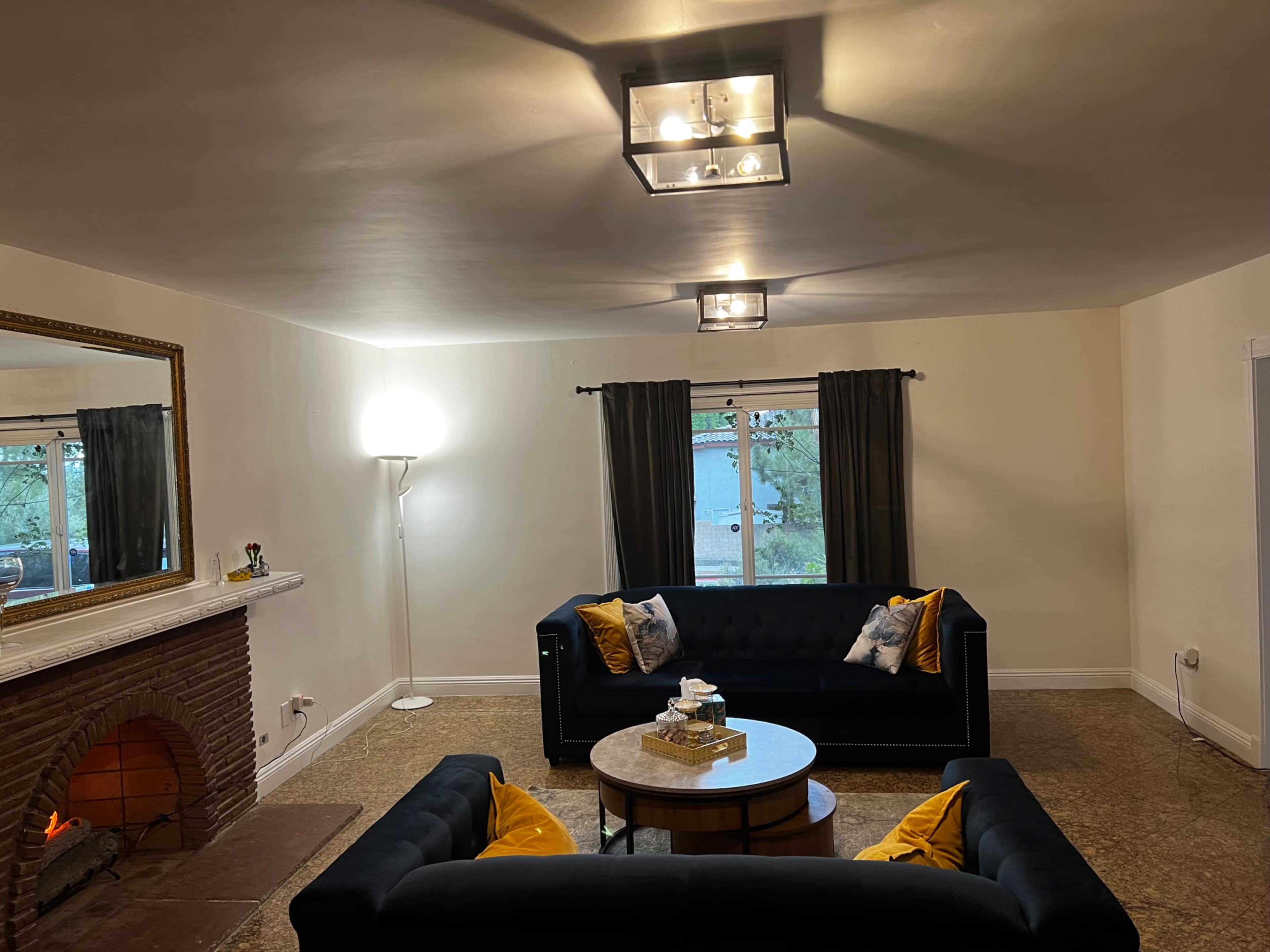 A living room features two black sofas adorned with yellow pillows, a round coffee table in the center, and a fireplace against one wall, with a lamp and windows providing natural light.
