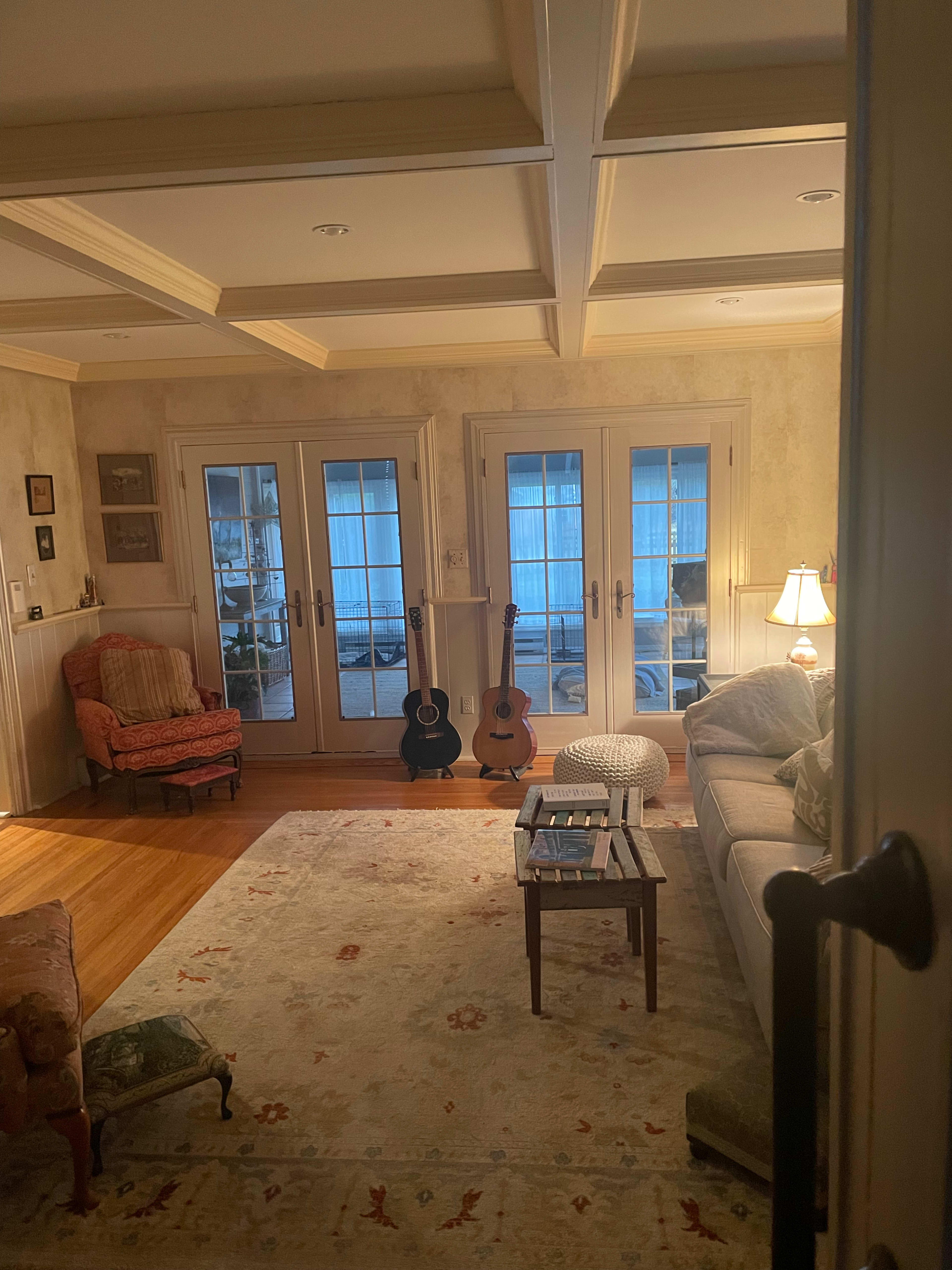 The image shows a cozy living room with two acoustic guitars leaning against a wall, a lighted lamp beside a sofa, and large glass doors leading to a patio.