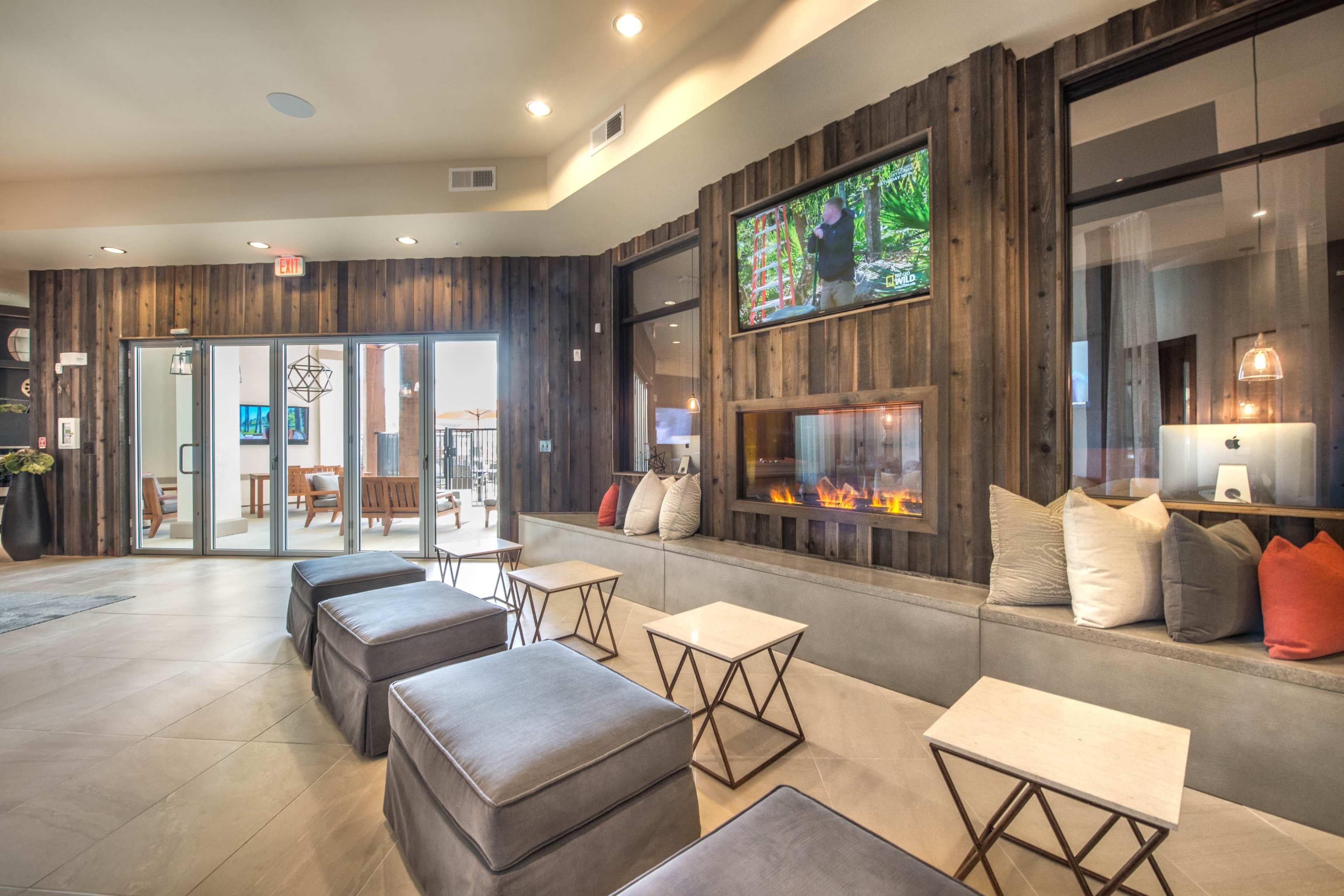 The image shows a contemporary lounge area featuring wooden walls, a modern fireplace, and seating with small tables.