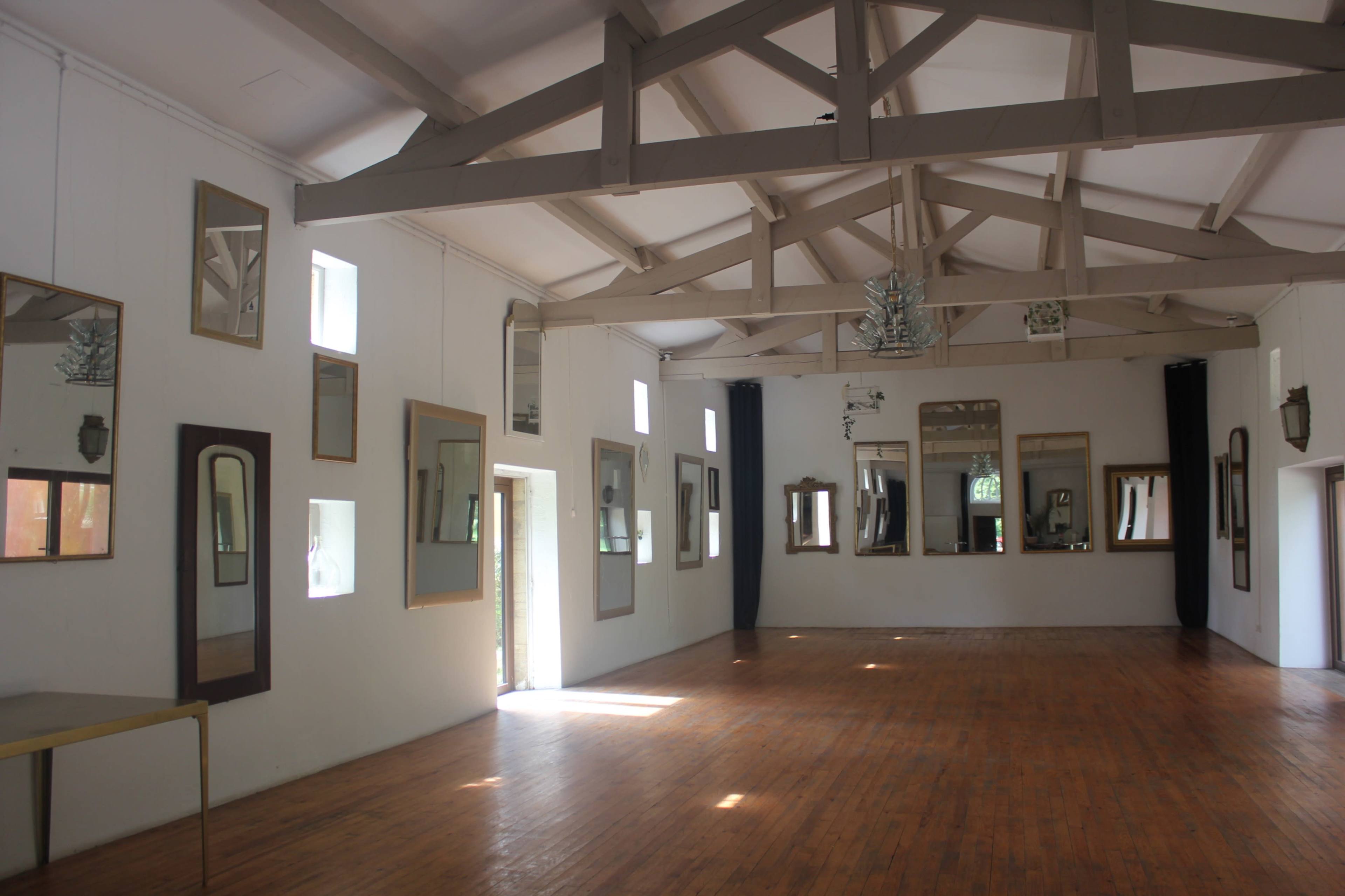 The interior of a spacious room features wooden flooring, several mirrors mounted on the white walls, and exposed wooden beams supporting the ceiling.