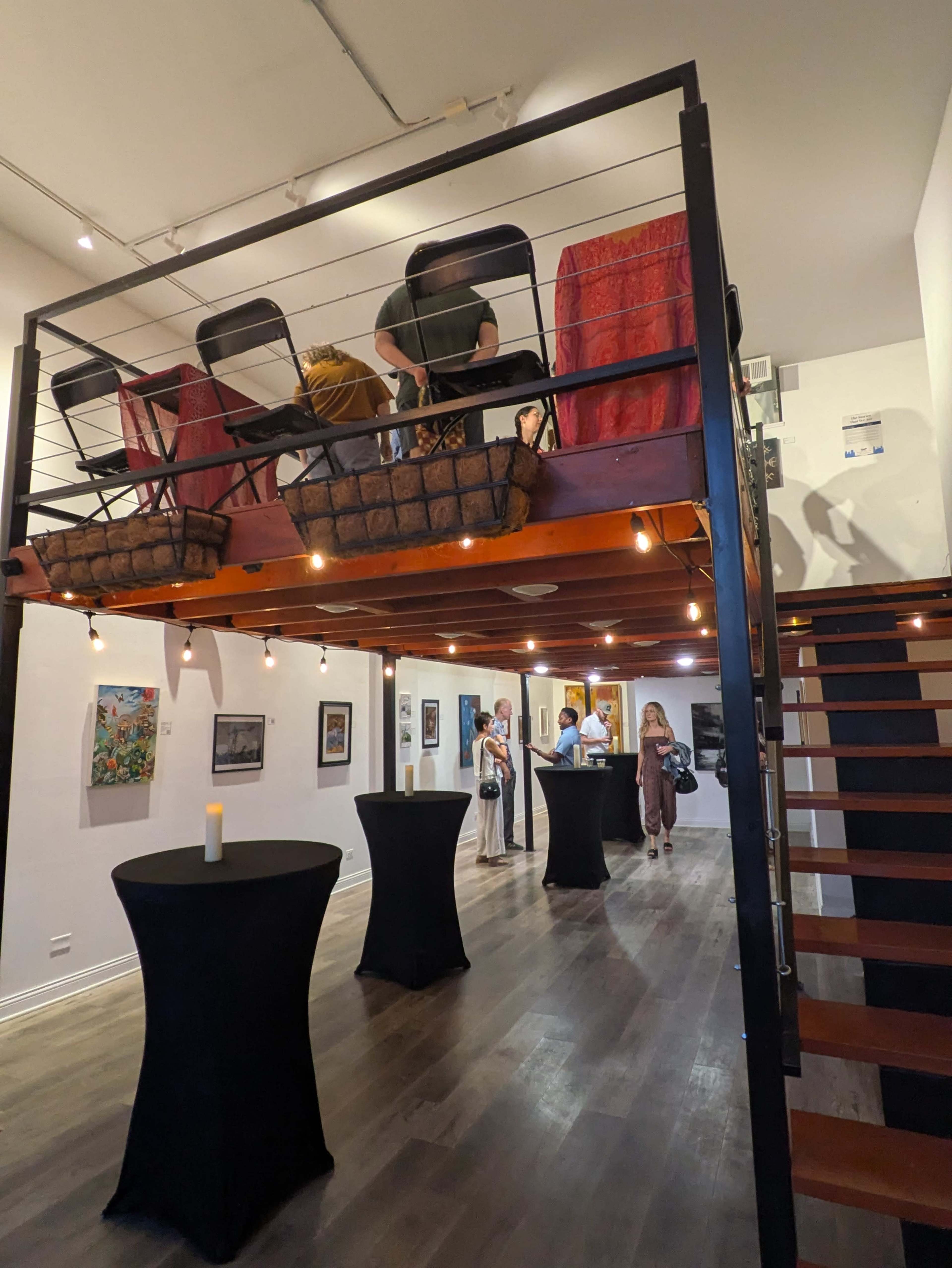 The image shows an art gallery with a lofted seating area above, featuring a staircase on the right and several people viewing artwork on the walls.