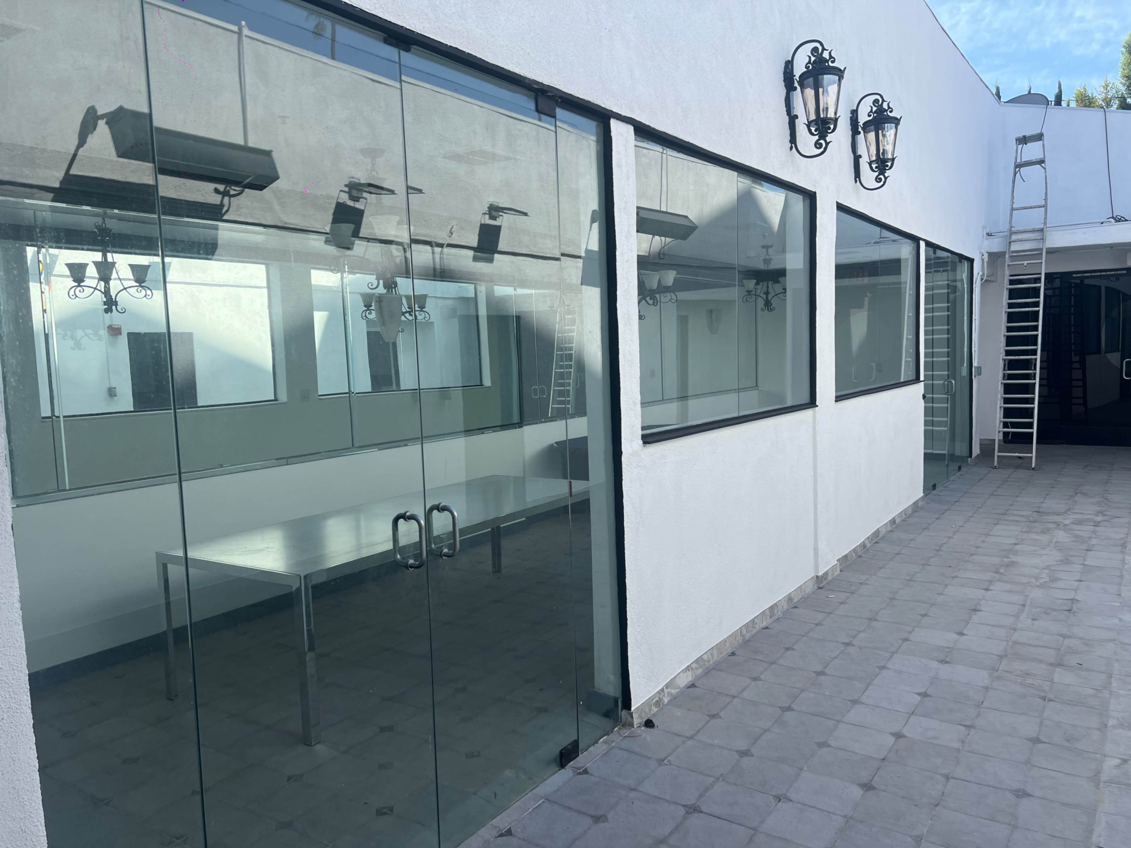 The image shows a modern building with large glass windows, a metal table inside, and an exterior wall featuring wall-mounted lanterns.