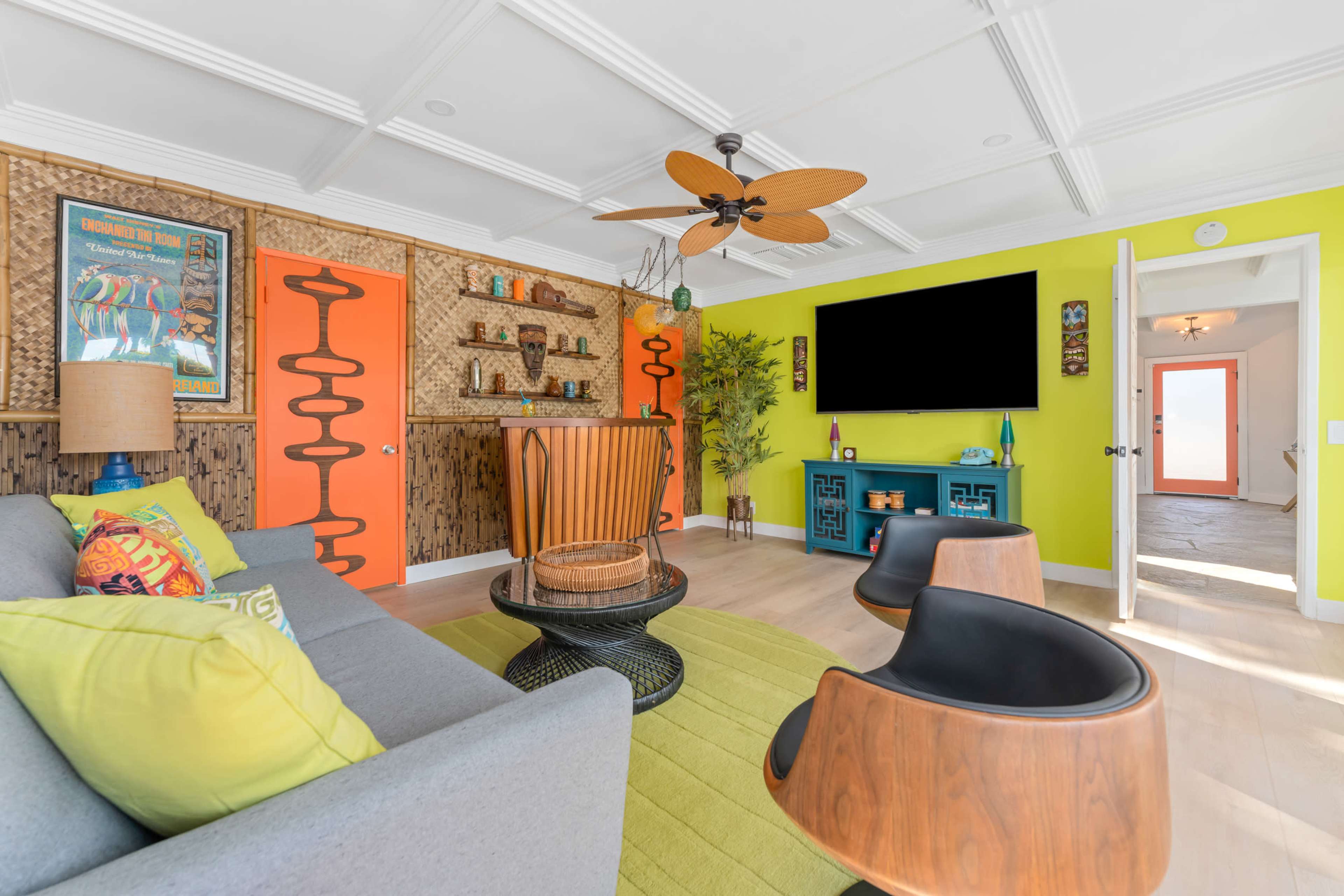 The living room features bright green and orange walls, a gray sofa, a wooden fan, and a large TV mounted on a blue sideboard.