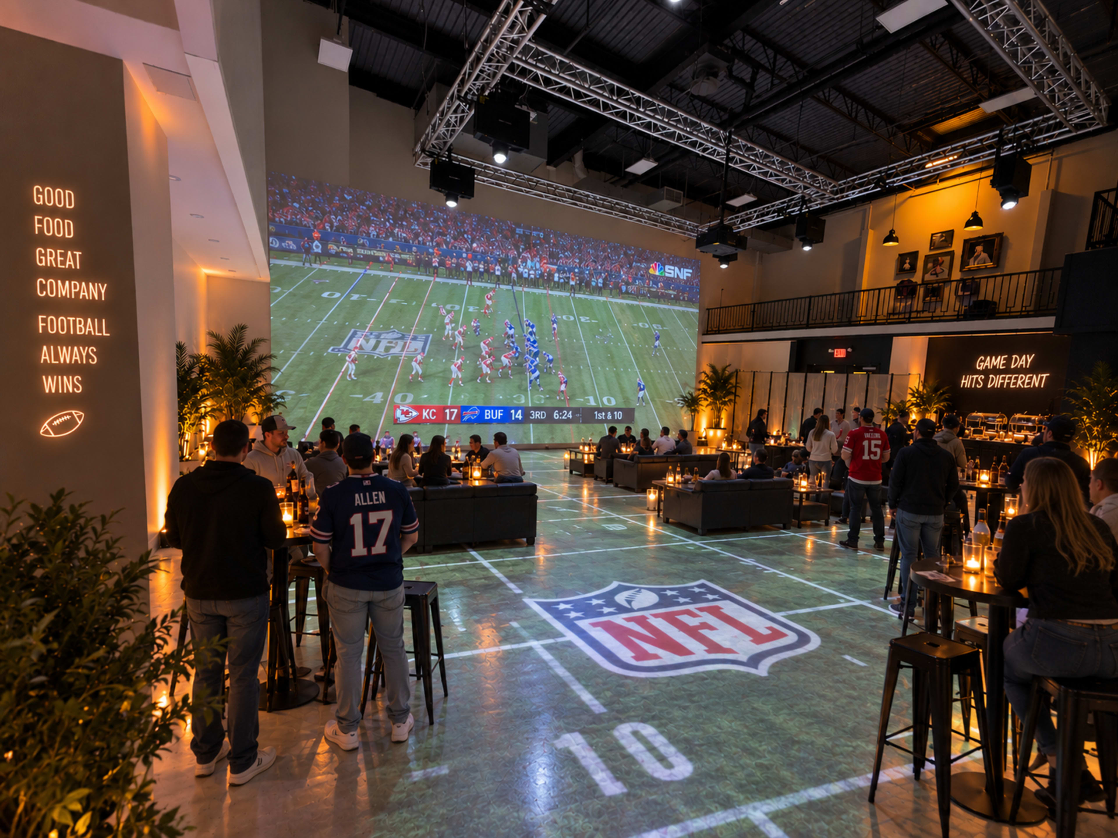 A sports bar features a large projected screen displaying a football game, with patrons seated at tables arranged around a football-themed floor design.