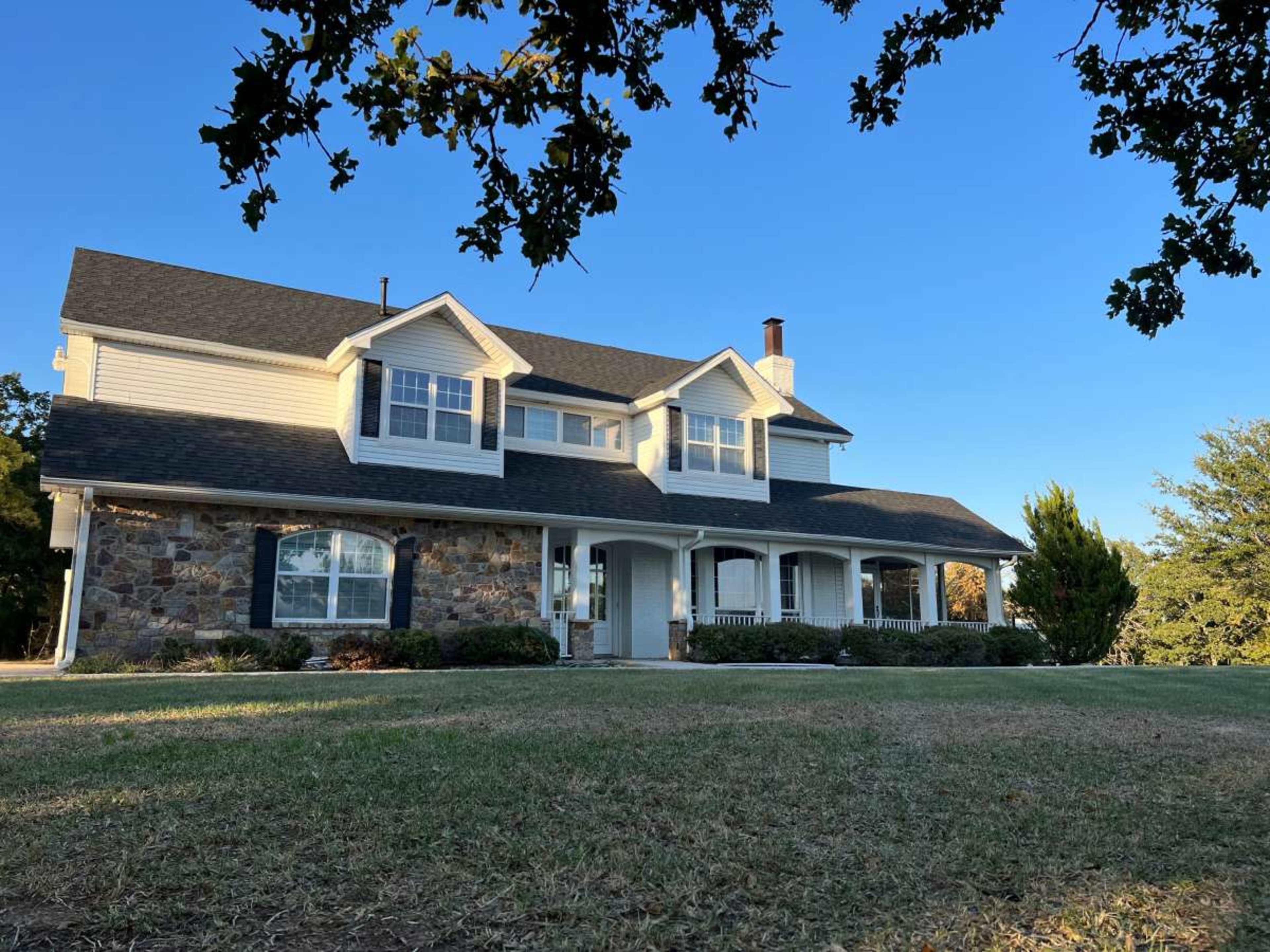 The image shows a two-story house with a stone and siding exterior, situated on a grassy lawn under a clear blue sky.
