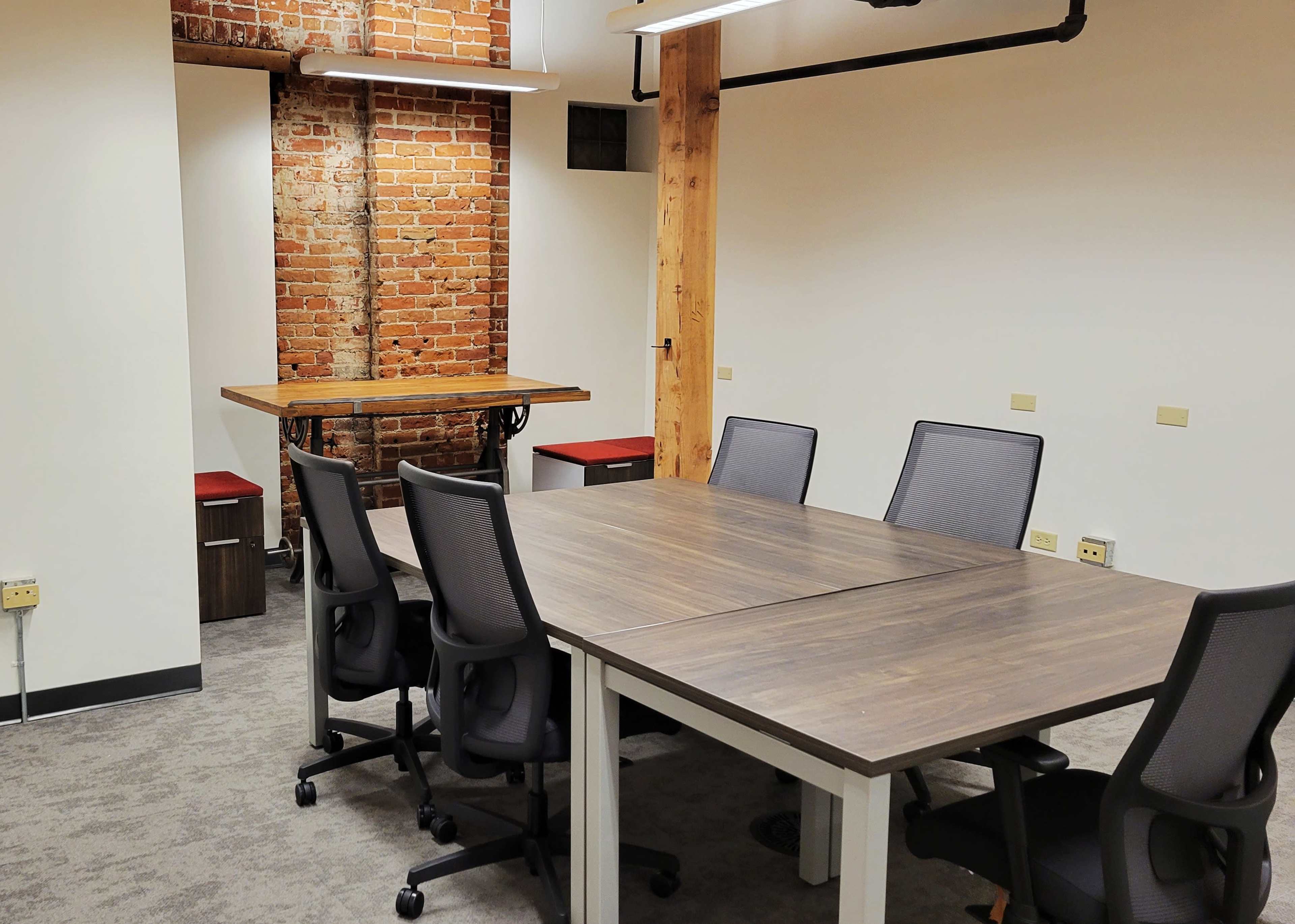 A small meeting room features a wooden table, four chairs, exposed brick walls, and modern lighting.