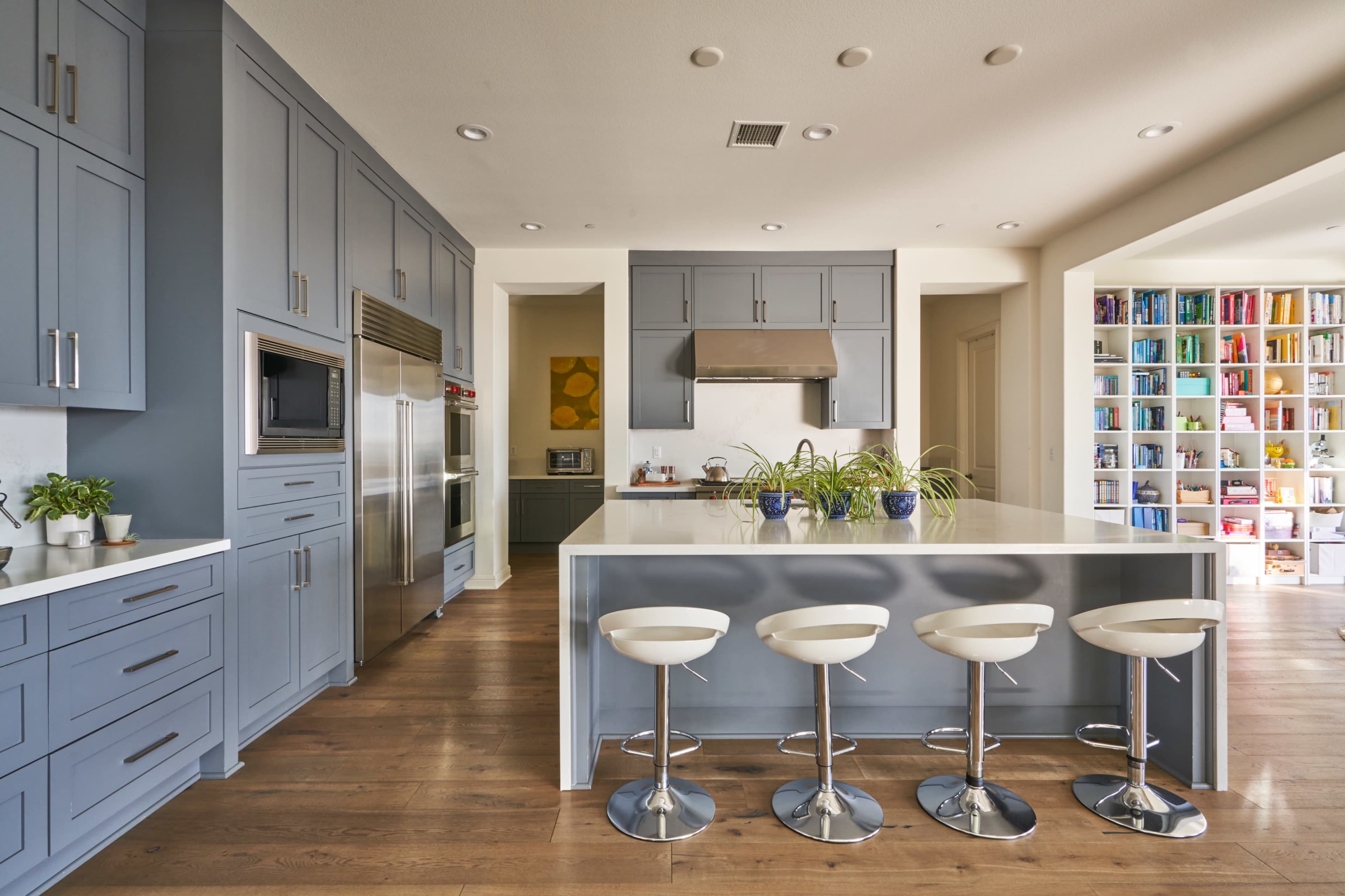 A modern kitchen features blue cabinetry, a central island with four white bar stools, and a pantry filled with colorful food items.