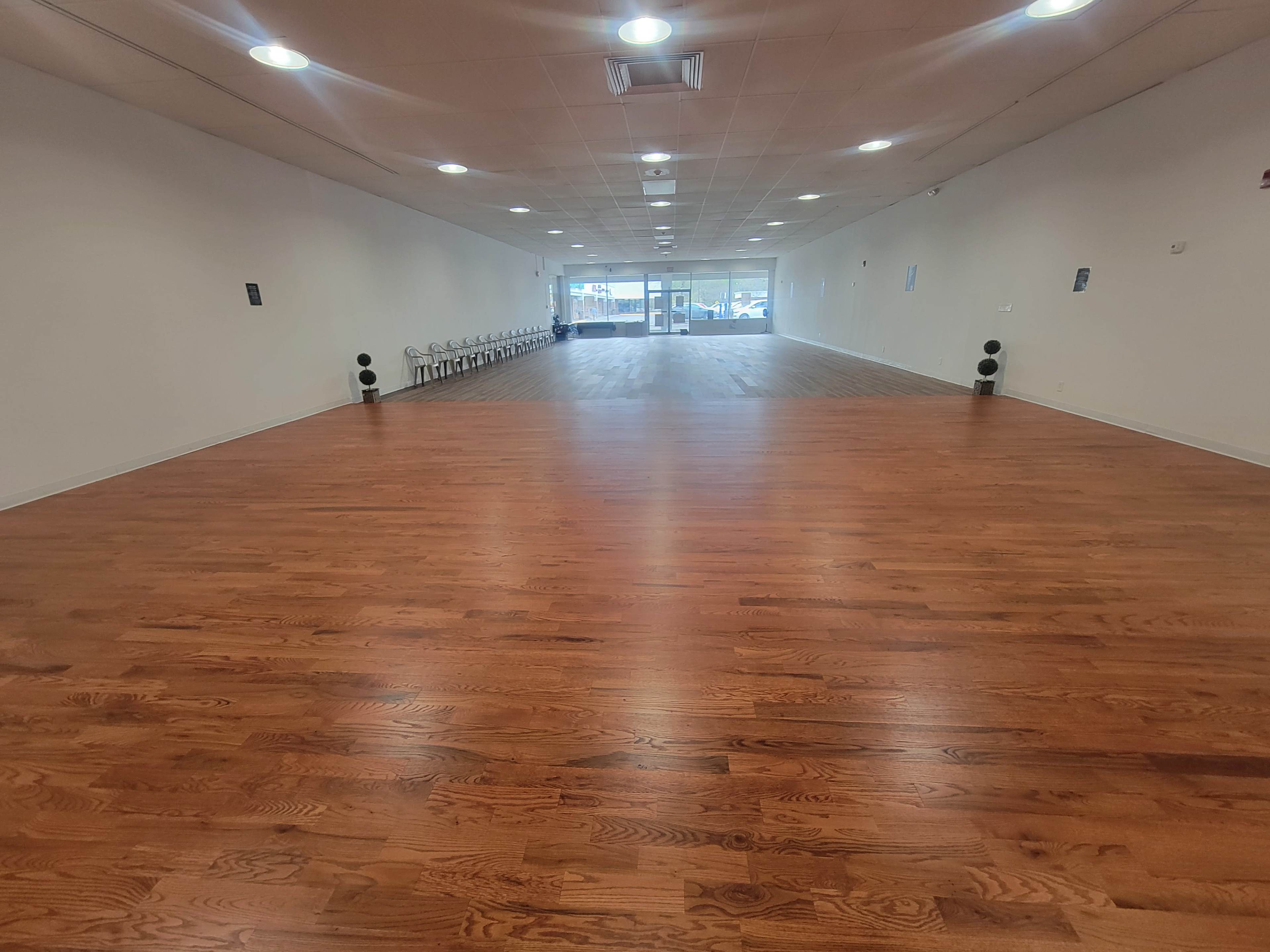 A spacious, empty room with polished wooden flooring and chairs lined up against one wall, leading to a glass entrance at the far end.