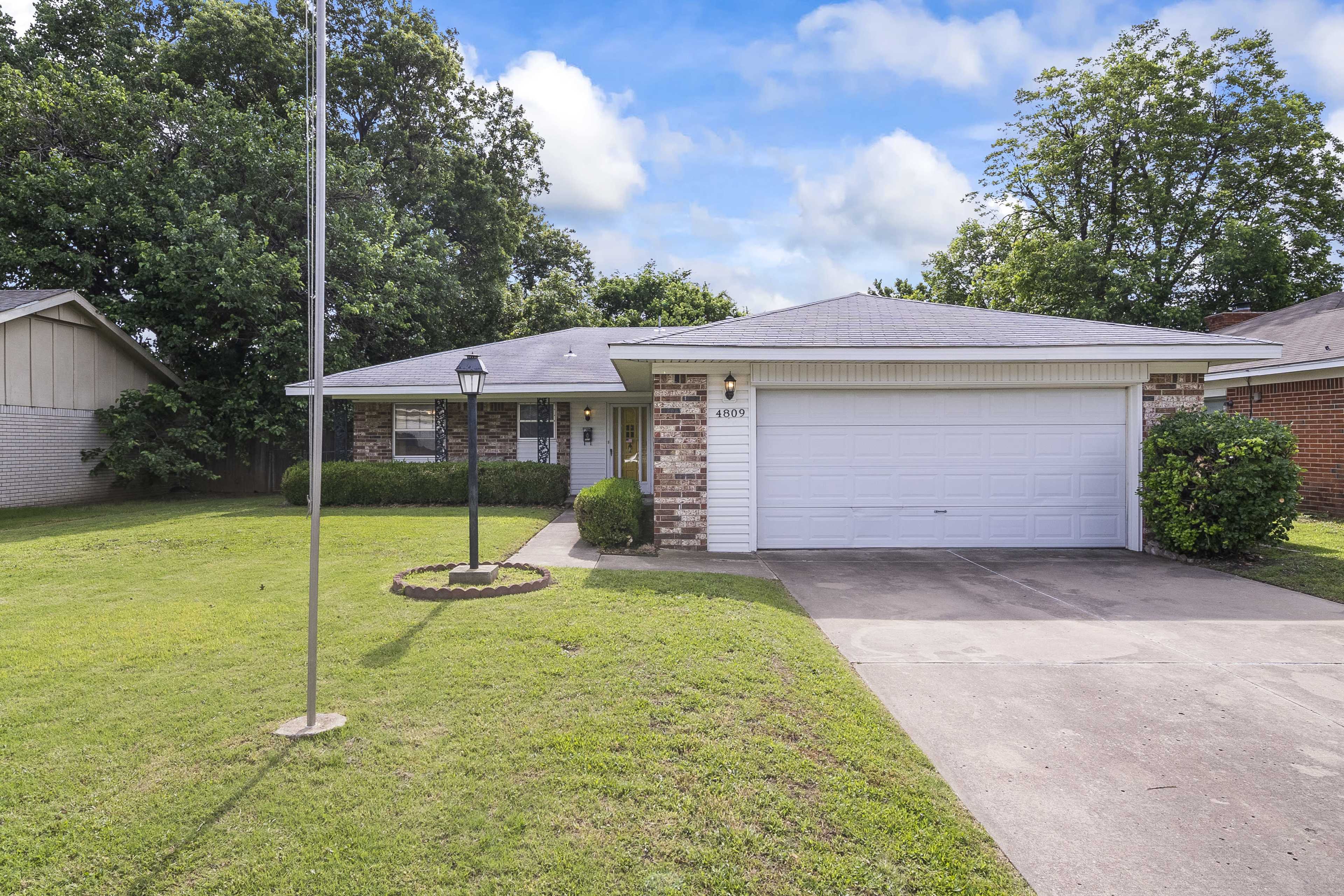 A single-story brick house with a two-car garage, surrounded by a lawn and a few trees.