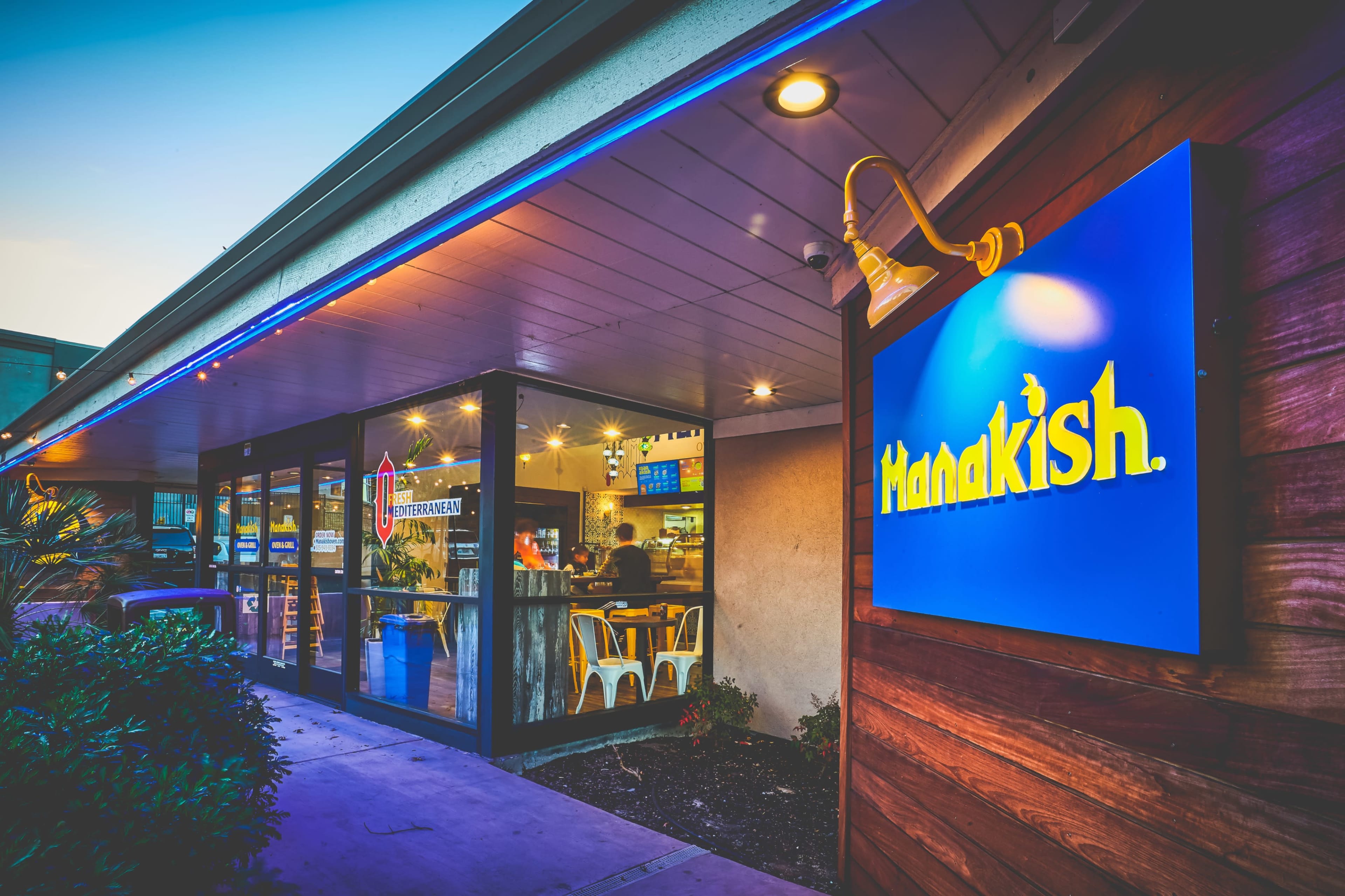 The image shows the exterior of a restaurant with a blue illuminated sign that reads "Manakish," featuring glass walls and a modern design.