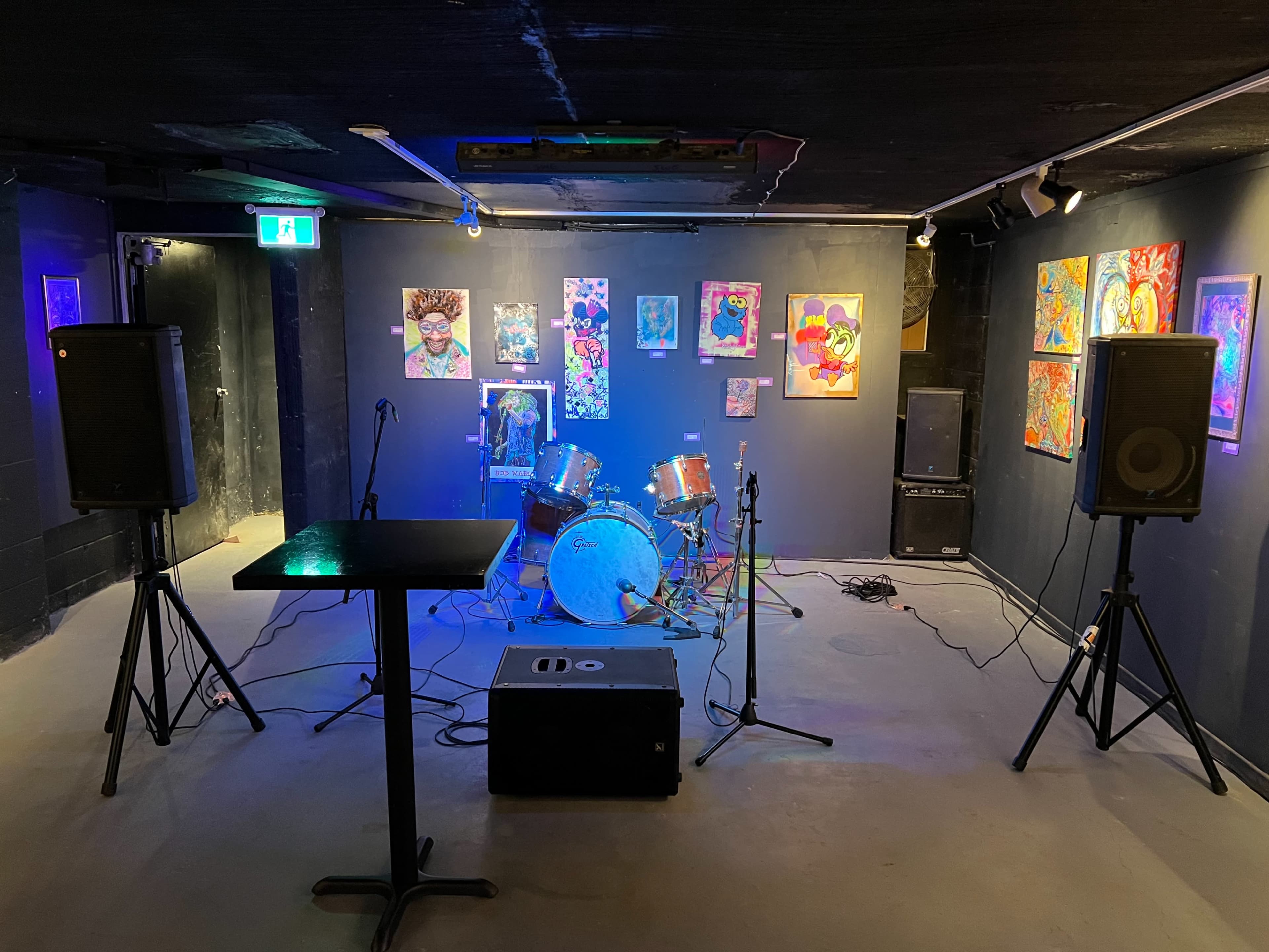 A music venue featuring a drum set and speakers, surrounded by colorful artwork on the walls.