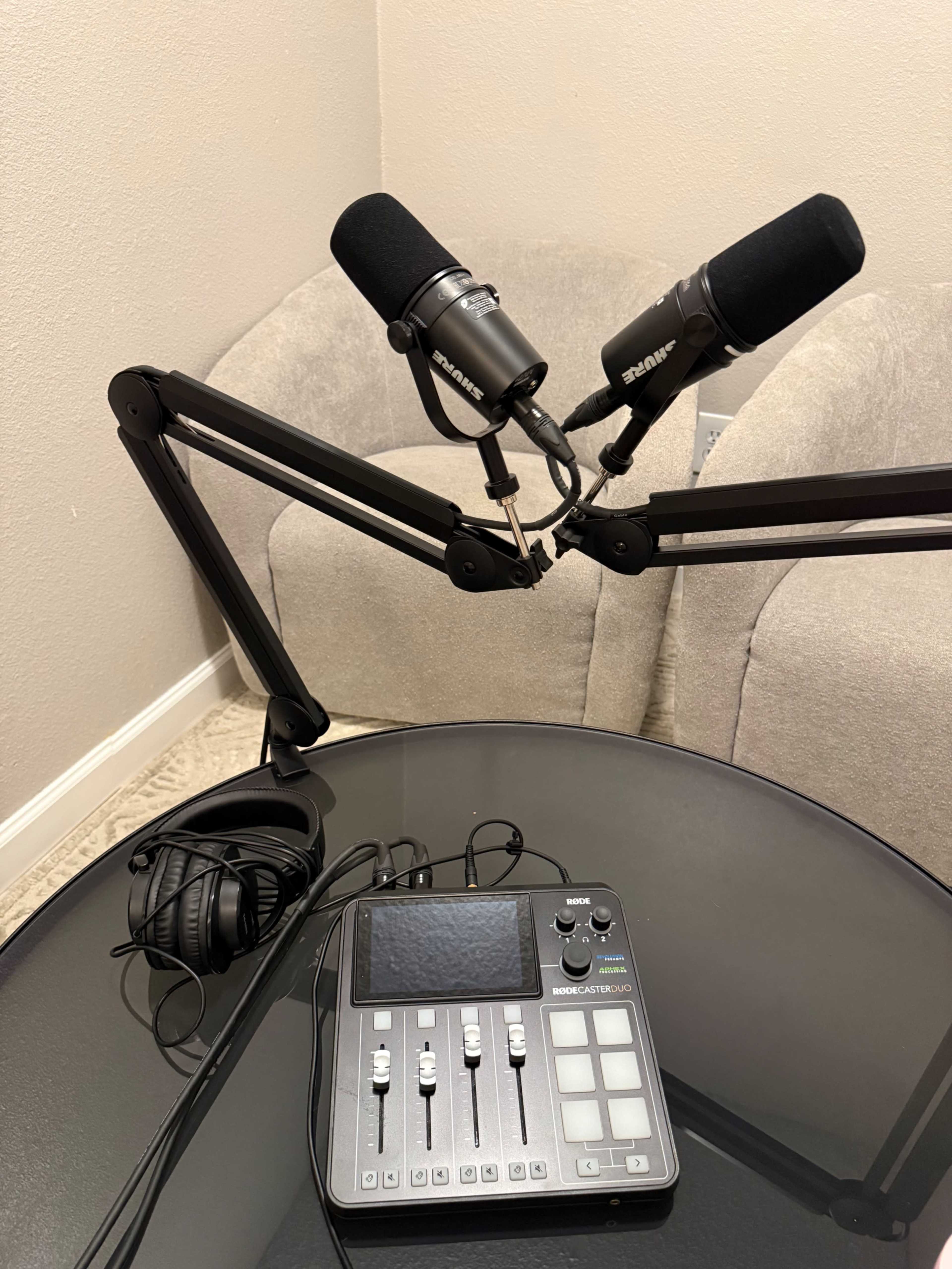 The image shows a podcasting setup with two microphones mounted on adjustable arms, a control panel, and a pair of headphones on a black table next to a beige chair.