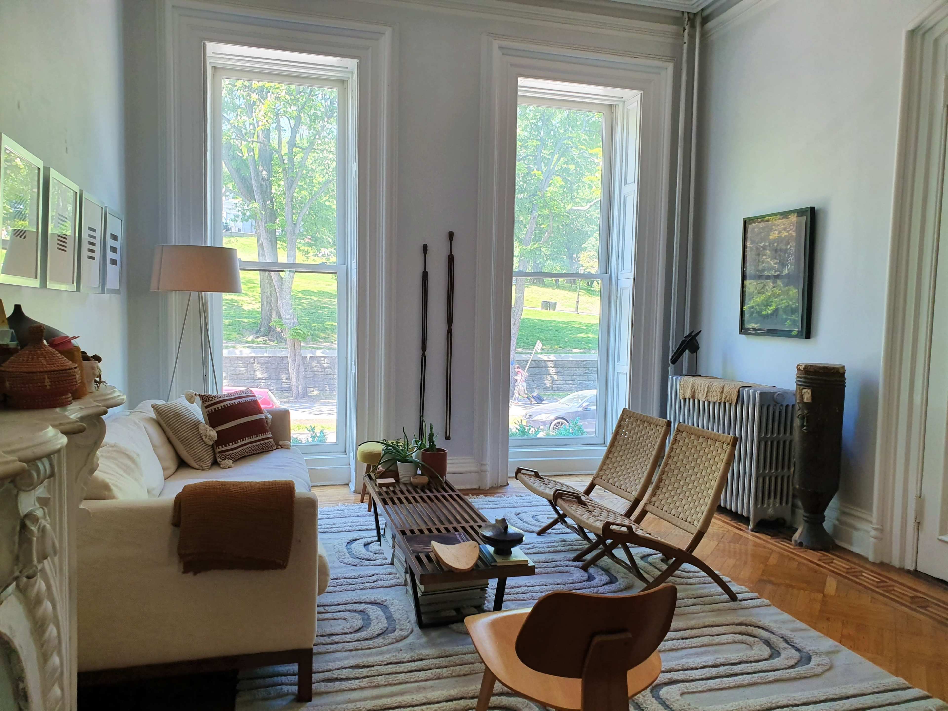 A bright living room features large windows, a white sofa, a wooden coffee table, two lounge chairs, and a radiator against the wall.