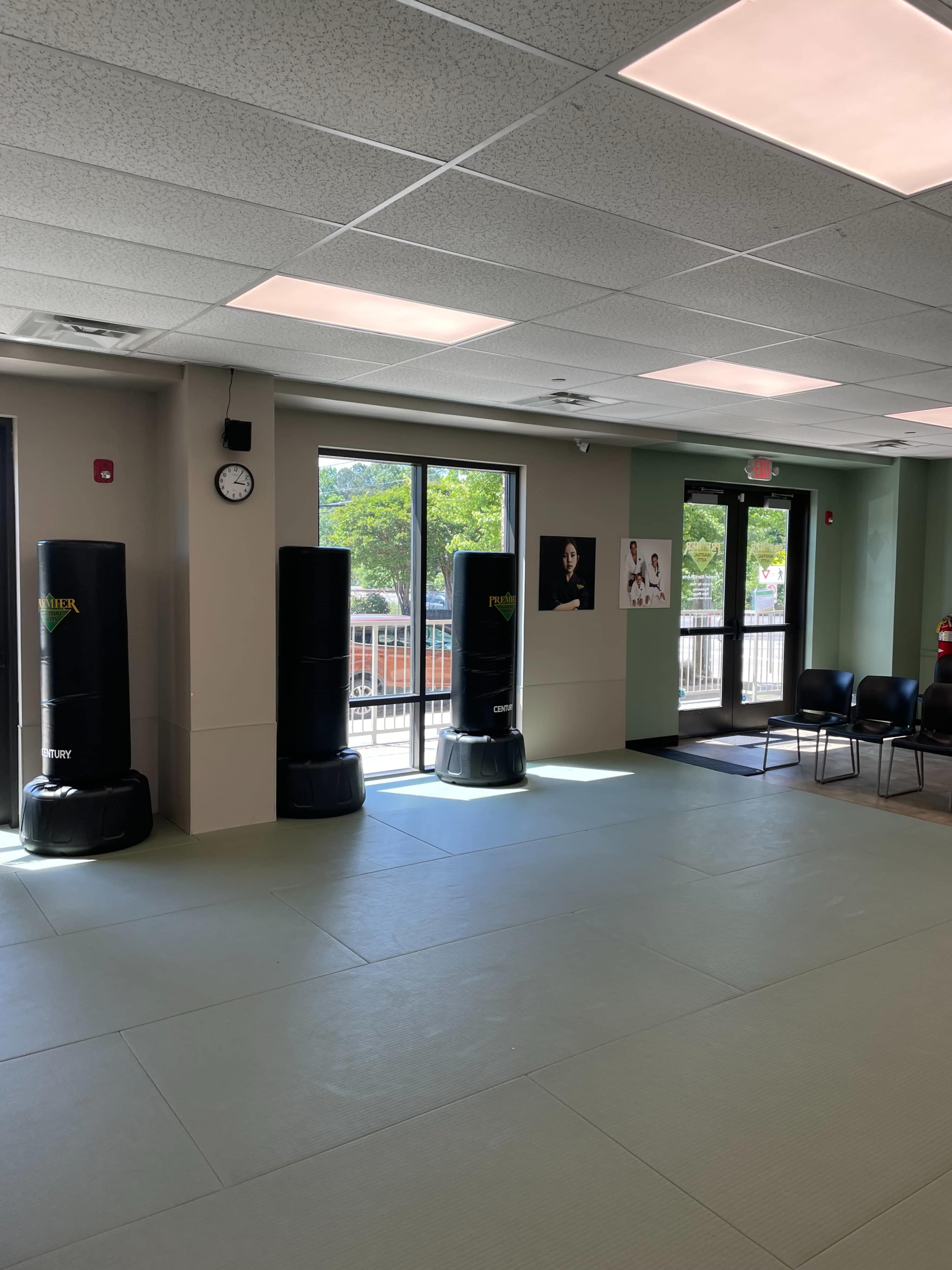 The image shows a well-lit martial arts studio with two black punching bags, a clock on the wall, and a row of chairs near large windows.