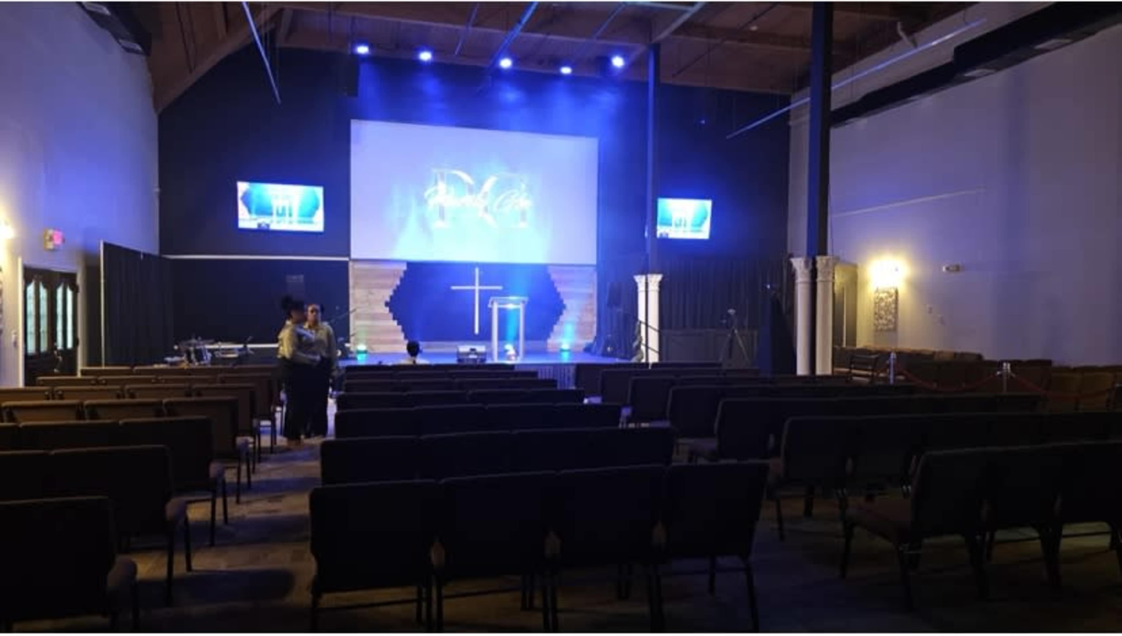 The interior of a church is shown, featuring rows of empty chairs facing a stage with a cross and illuminated screens.