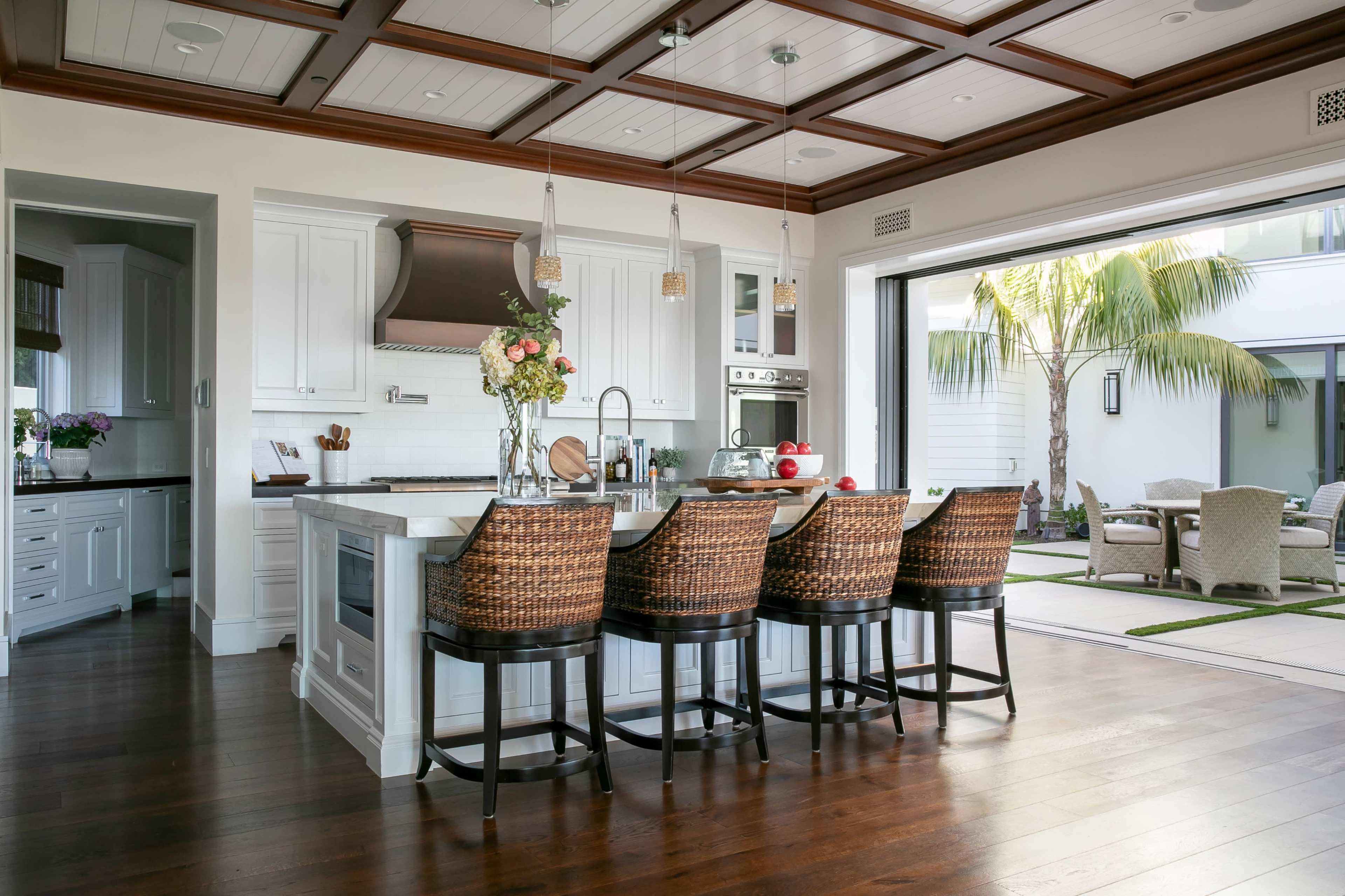 The image shows a modern kitchen with a large island, bar seating, and an open view to a patio featuring palm trees.