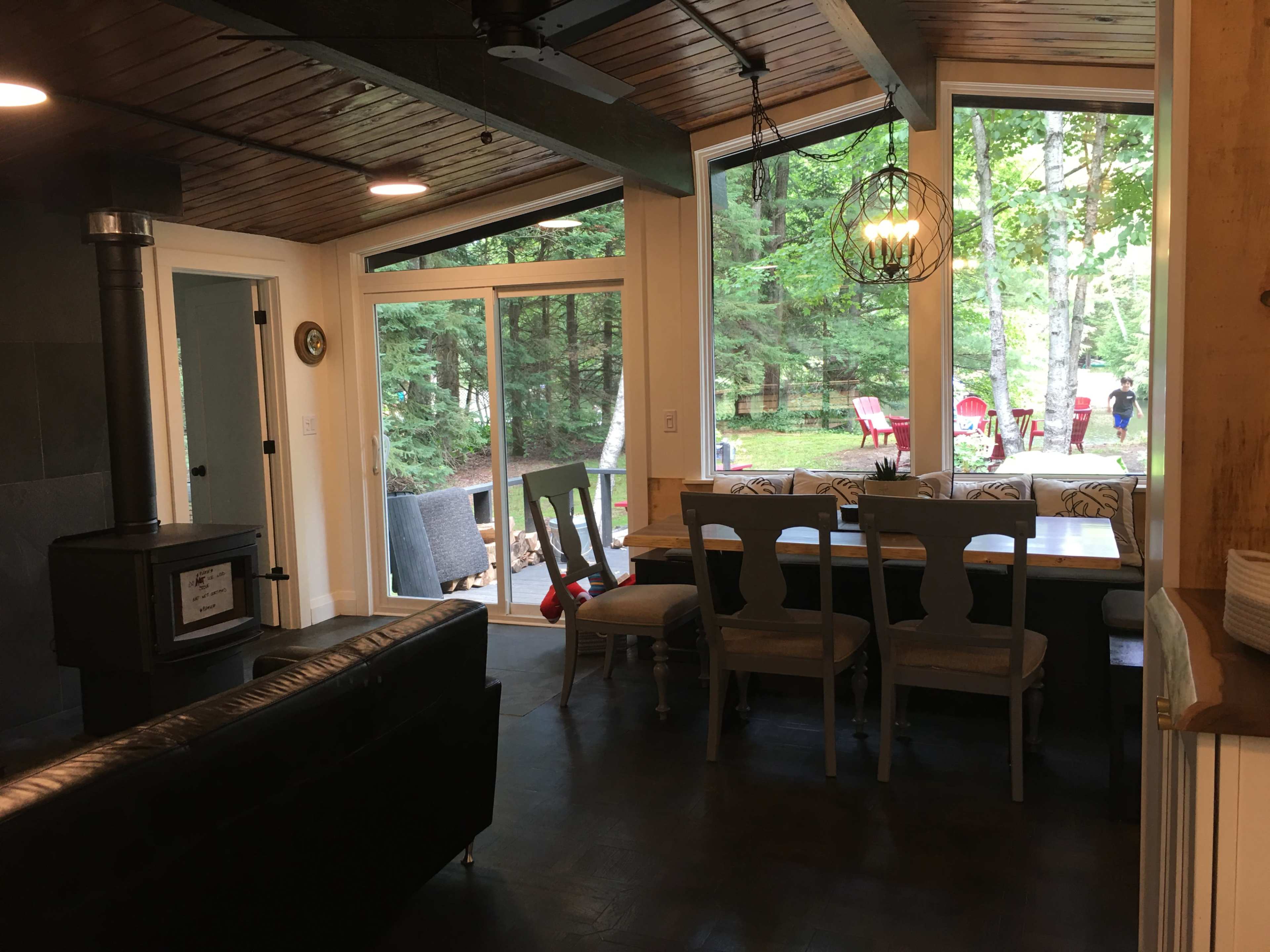 A cozy kitchen-dining area with a wooden table surrounded by chairs, adjacent to a large window overlooking a view of green trees and outdoor seating.