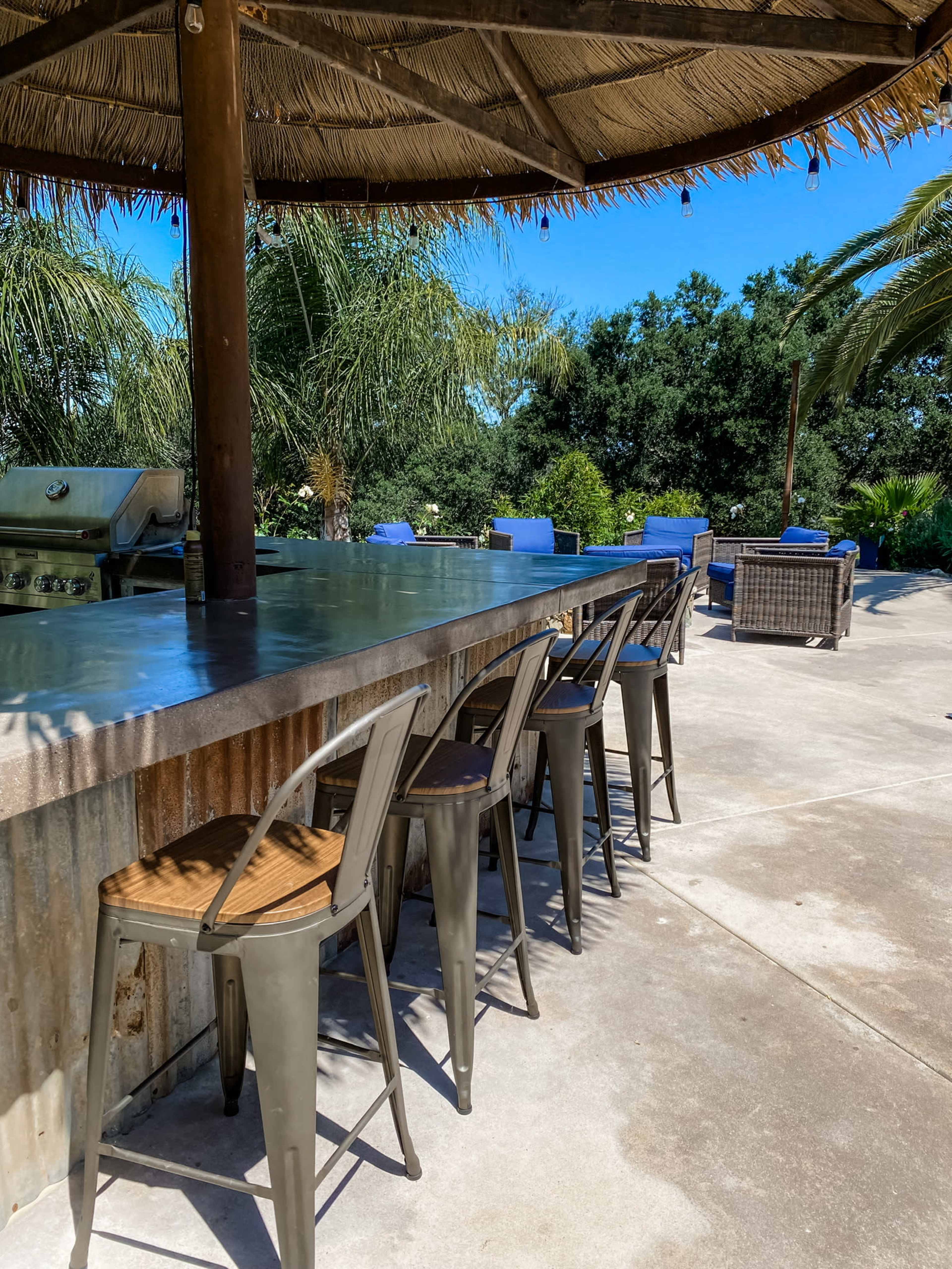 An outdoor bar with metal stools and a thatched roof is set against a backdrop of palm trees and lounge chairs by the pool.