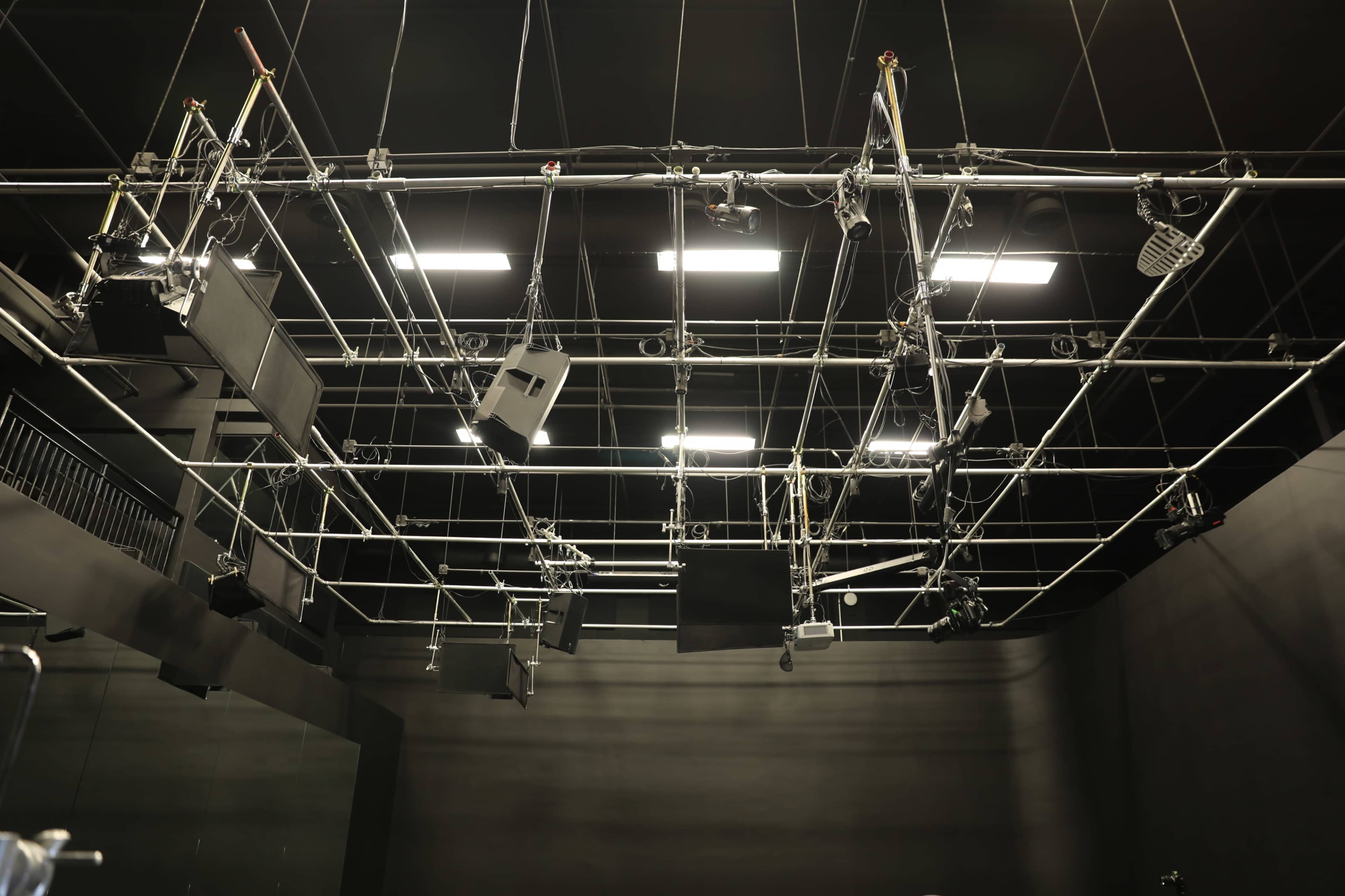 The image shows a grid-like structure of lighting and audio equipment suspended from the ceiling of a dark studio space.