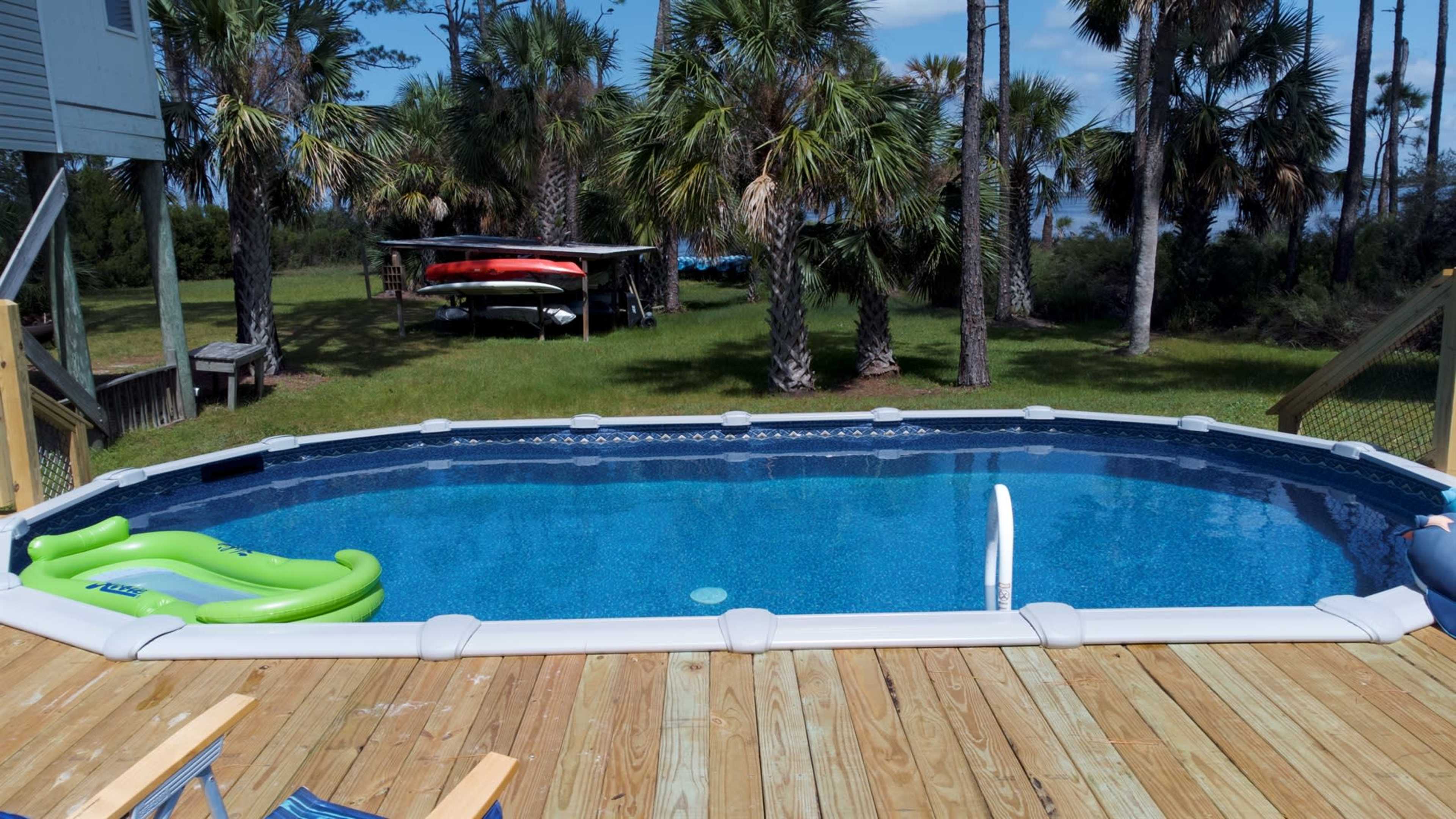 A blue swimming pool surrounded by palm trees and a grassy area, with kayaks stored on a rack in the background.