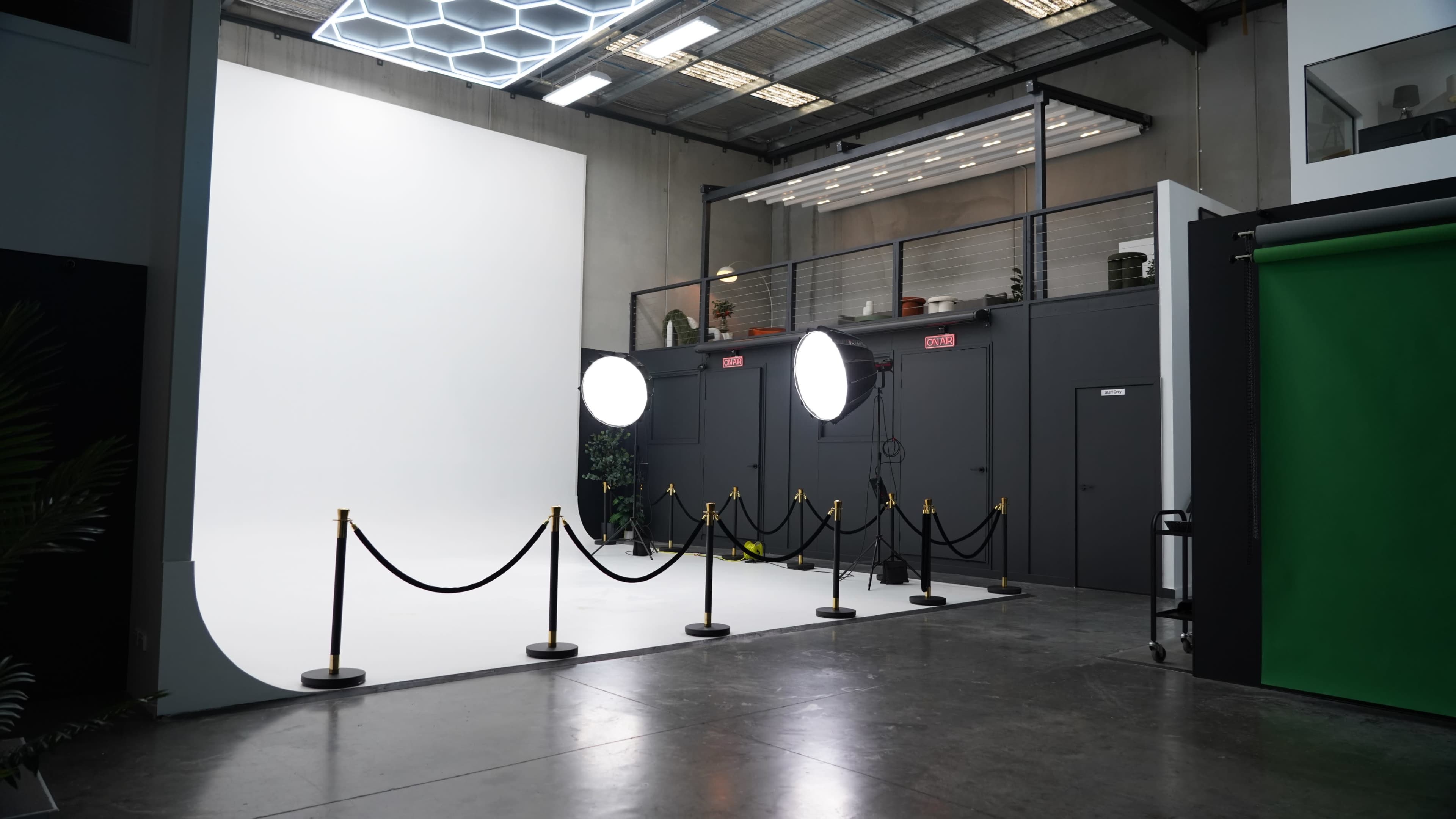 The image shows a photography studio featuring a white backdrop, two large softbox lights, and areas partitioned by black ropes, with a green screen visible on one side.