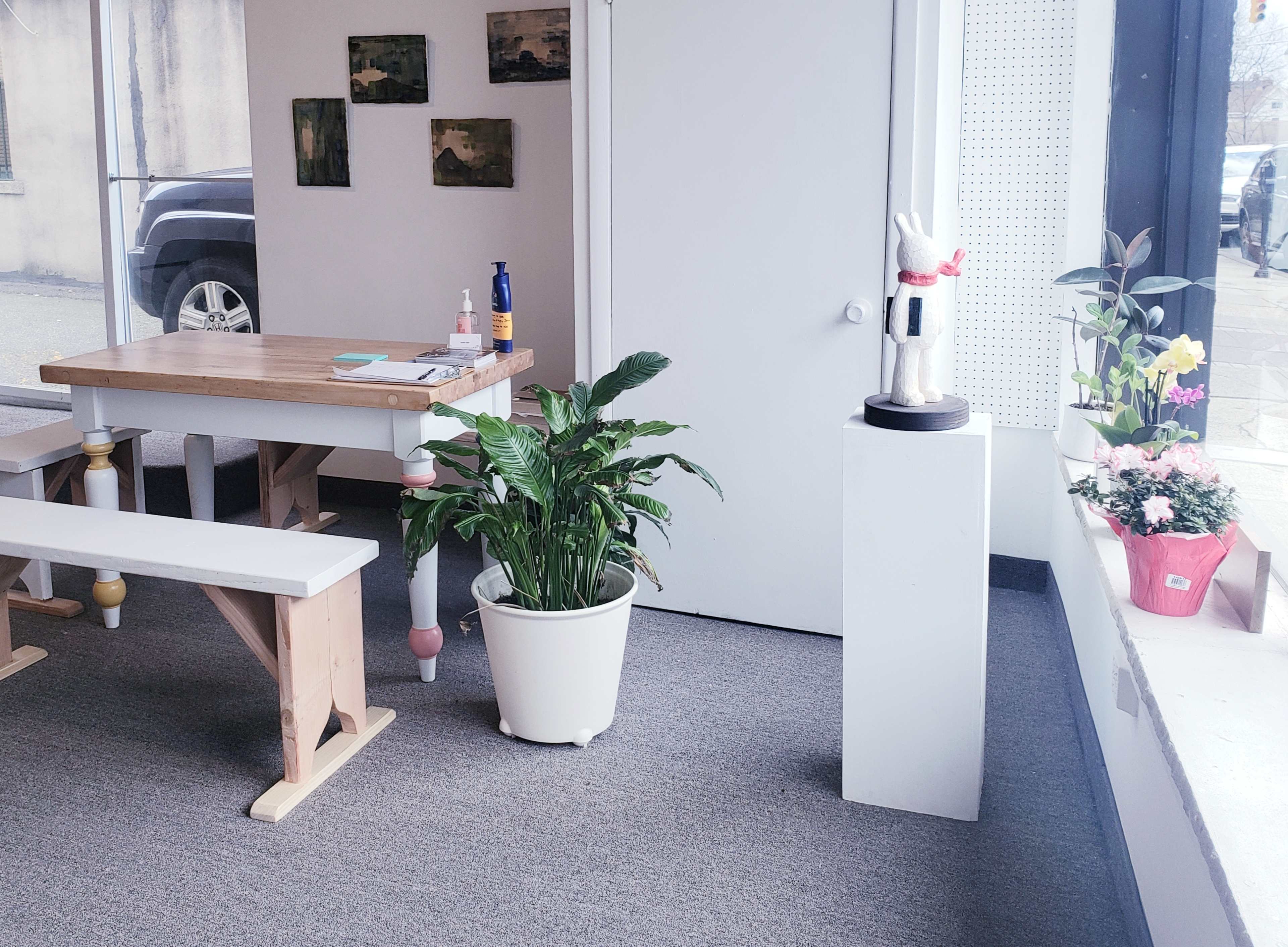 A spacious, brightly lit room features a wooden table and benches, a large potted plant, and decorative sculptures next to flowering pots by a window.