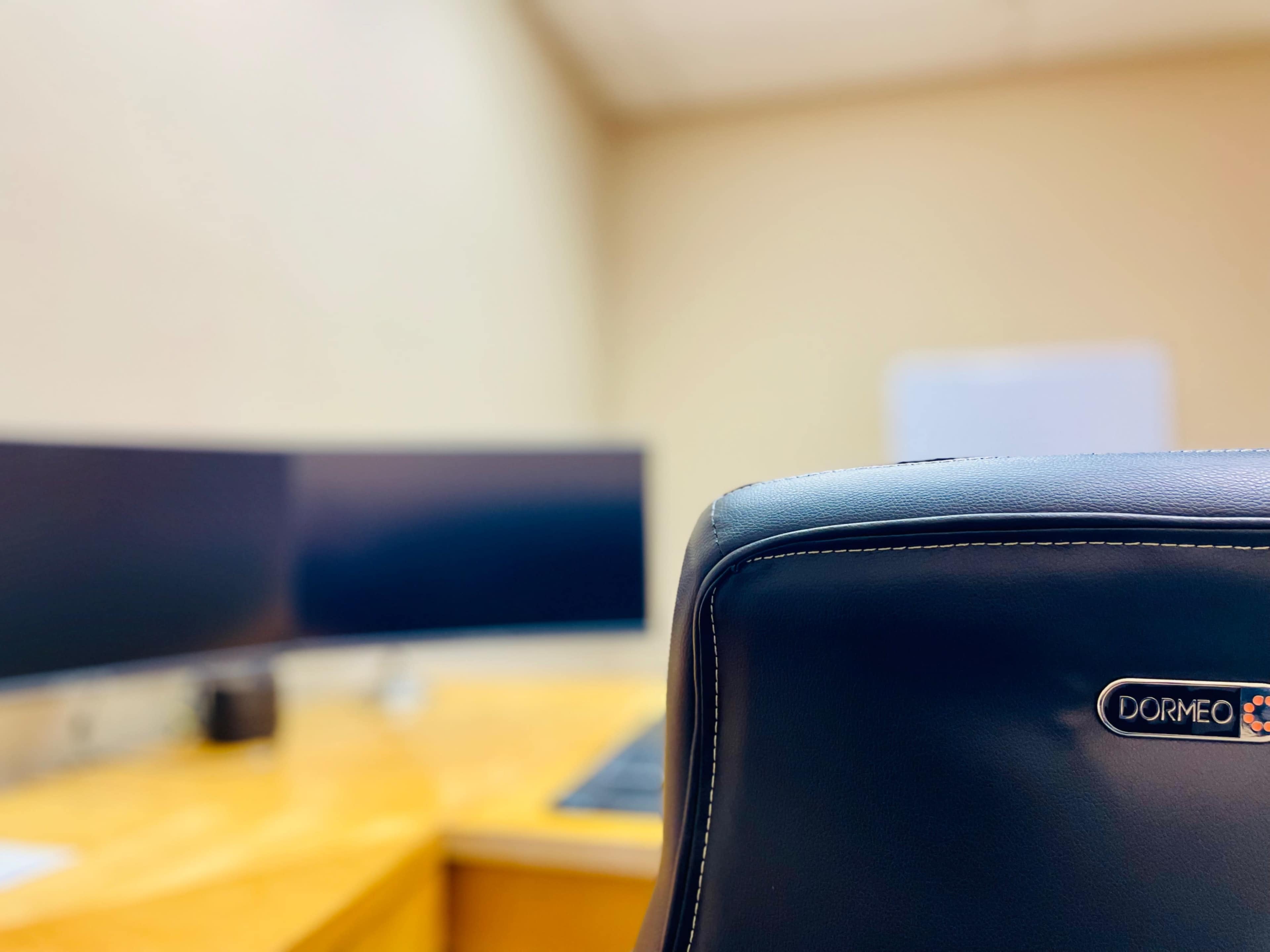 A black ergonomic office chair is positioned in front of a wooden desk with dual monitors in a beige office environment.