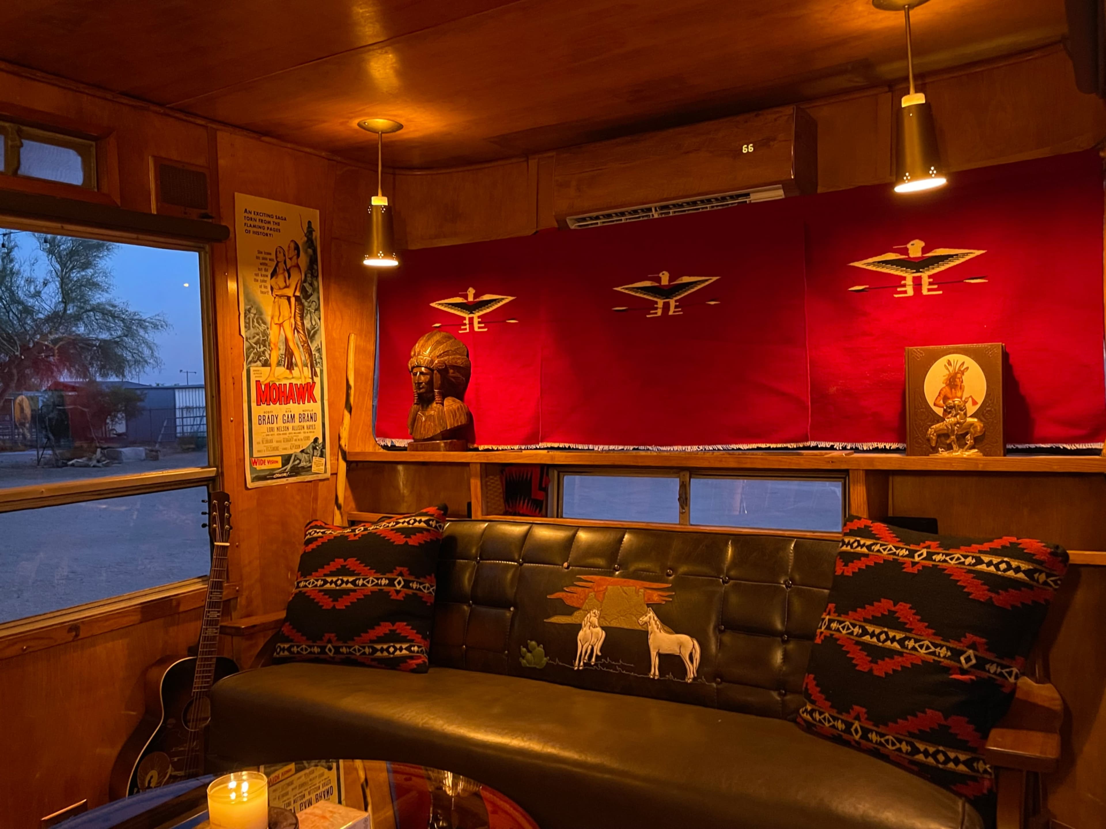 The interior of a vintage trailer features a black leather bench, patterned throw pillows, and decorative items including a wooden sculpture and retro posters.
