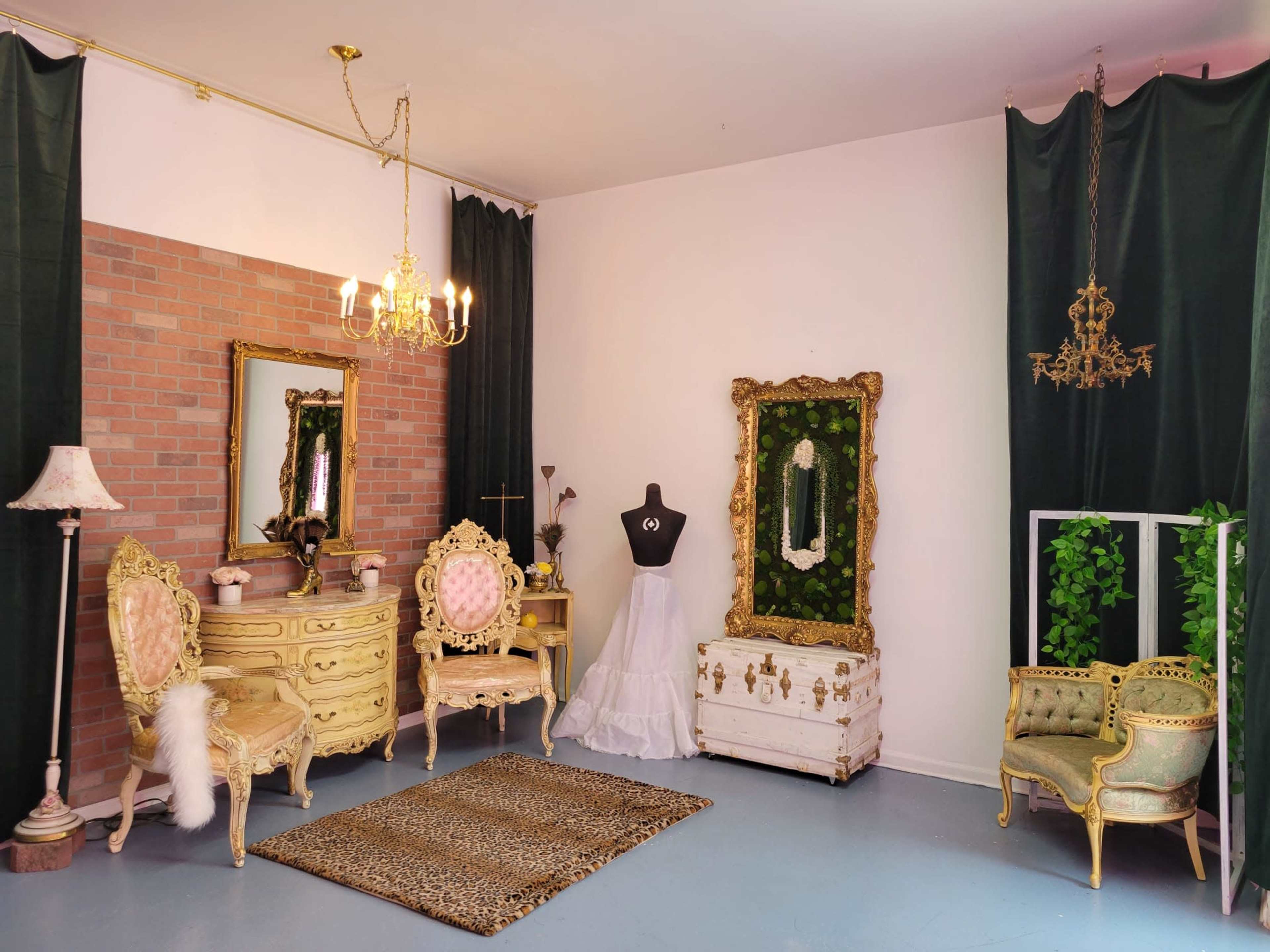 An elegant room featuring vintage furniture, a decorative mirror, a dress form, and greenery, arranged on a light-colored floor.
