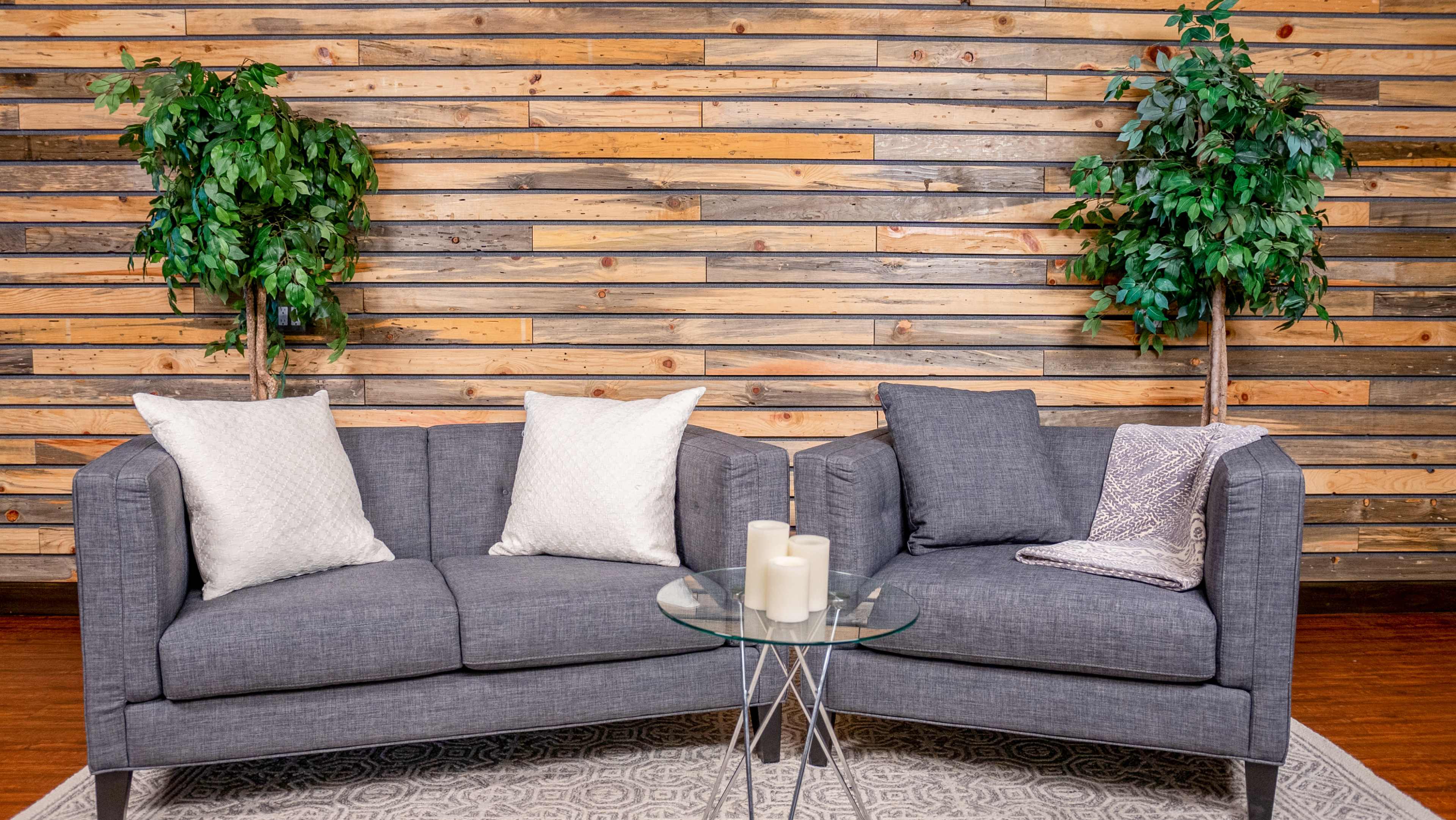 A gray sectional sofa with two decorative pillows and a small glass coffee table is placed against a wooden paneled wall, flanked by two potted plants.