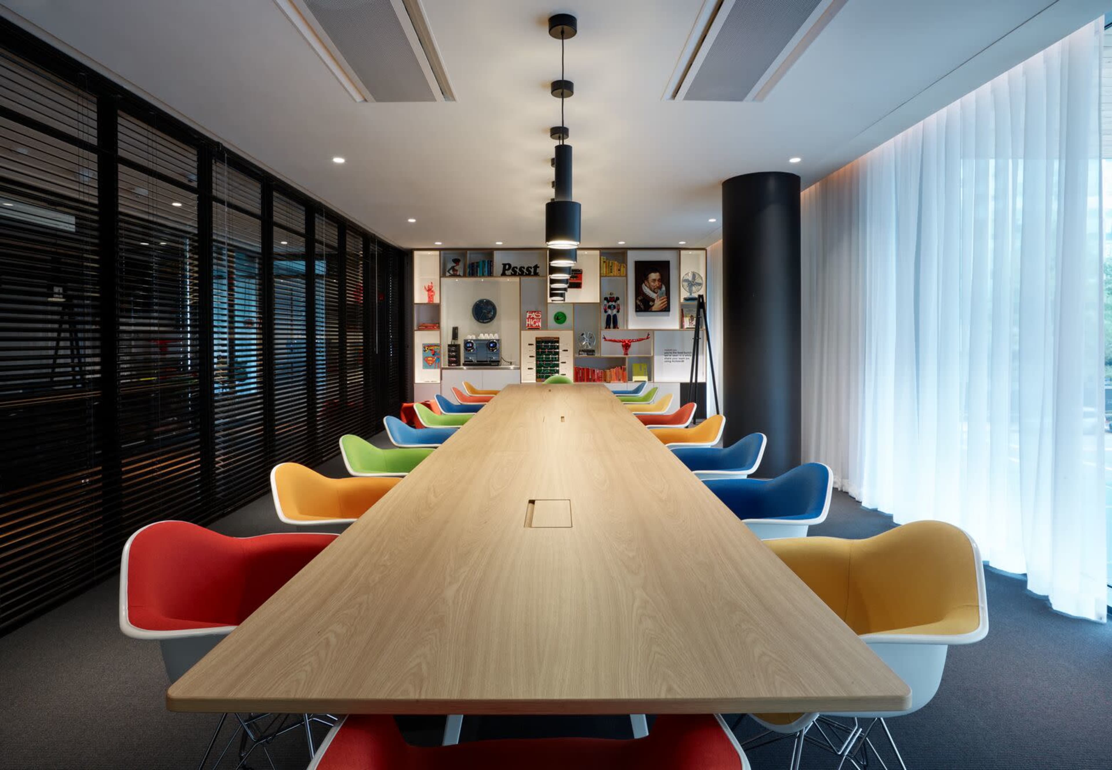 A long wooden table is surrounded by colorful chairs in a modern conference room with large windows and shelves displaying books and decor.