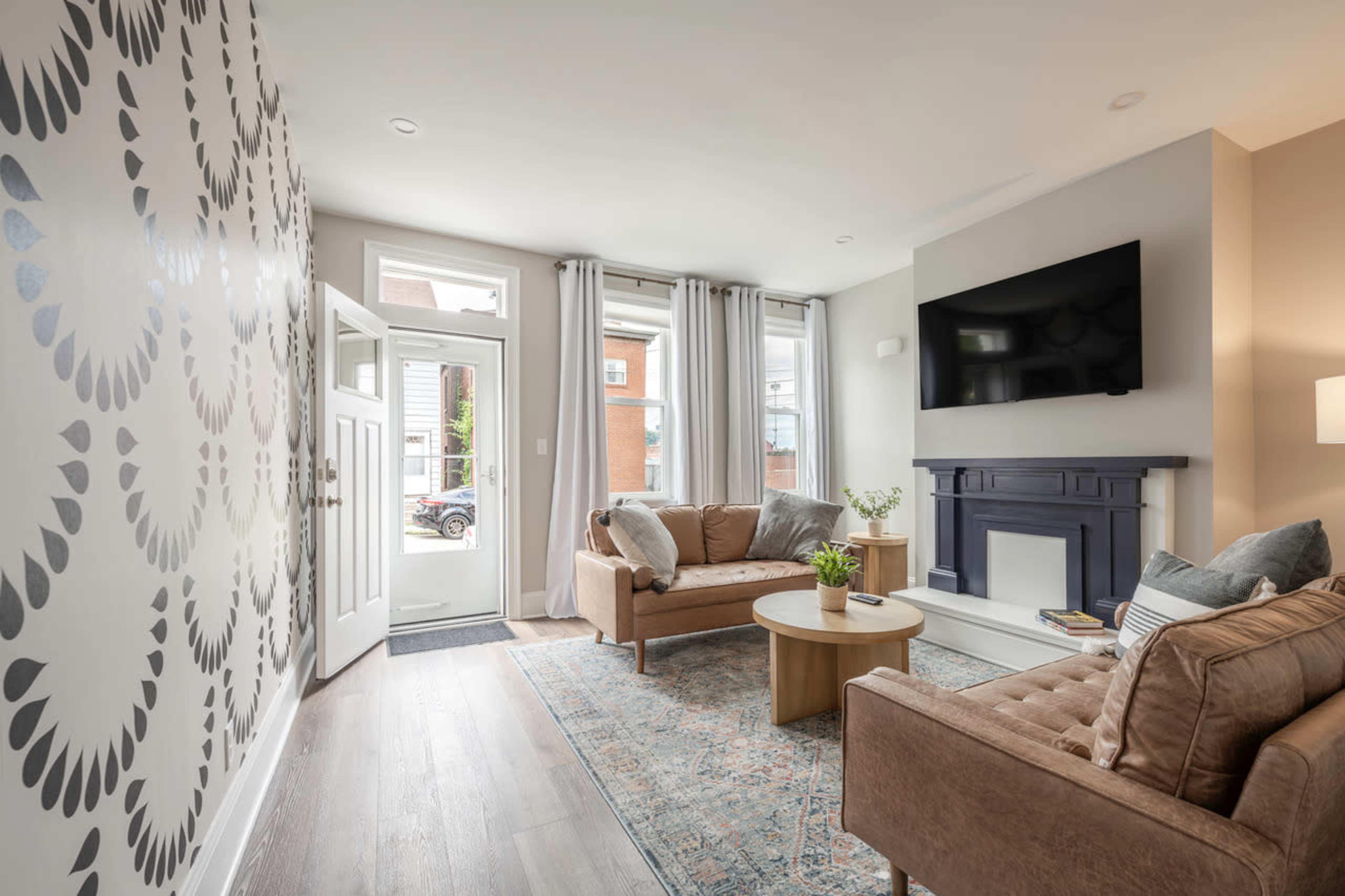 The image shows a modern living room featuring beige sofas, a round wooden coffee table, a flat-screen TV mounted on the wall, and large windows with sheer curtains.
