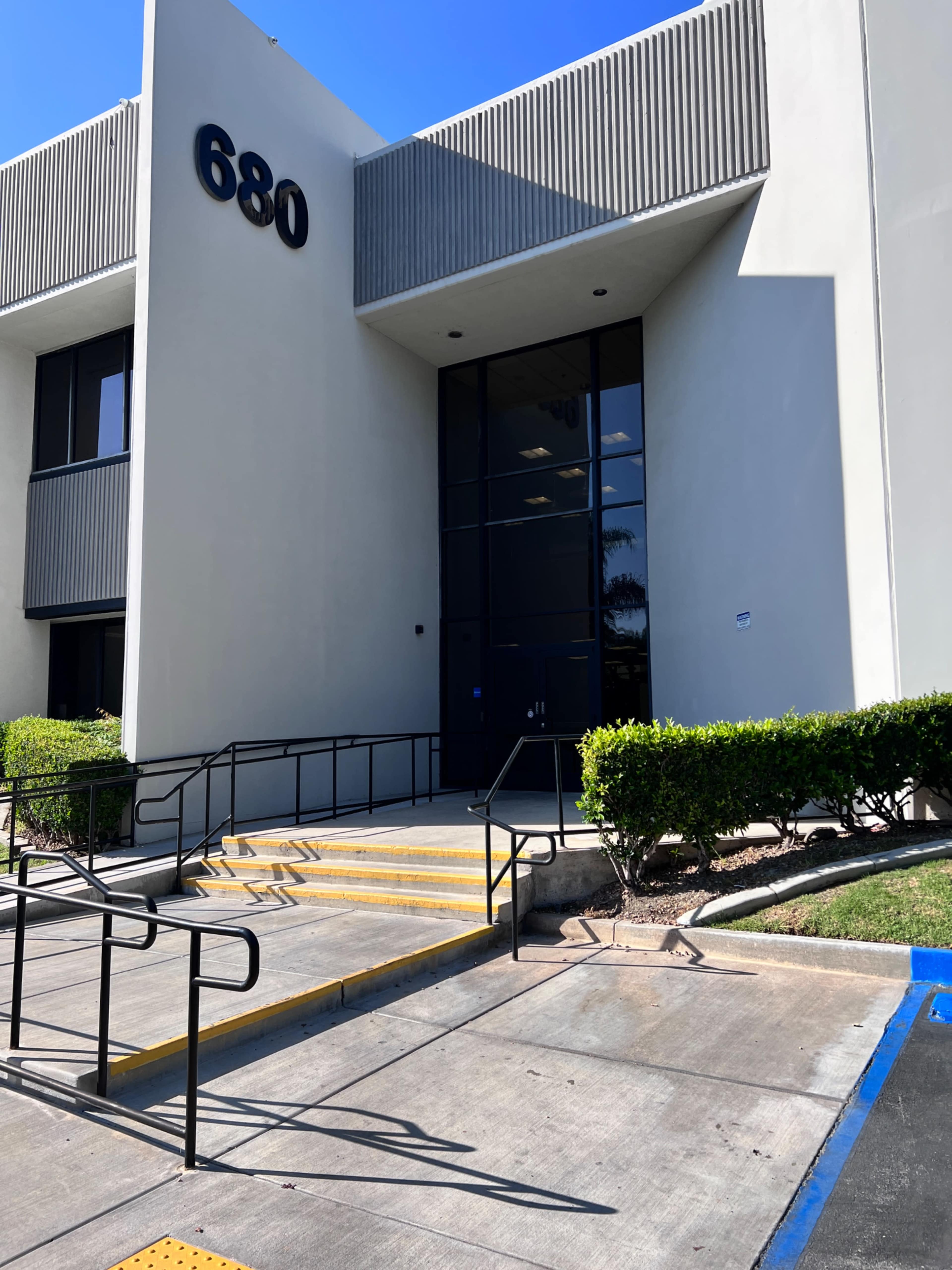 The entrance of a modern building with the number 680 displayed on the facade, featuring a set of stairs and a landscaped area.