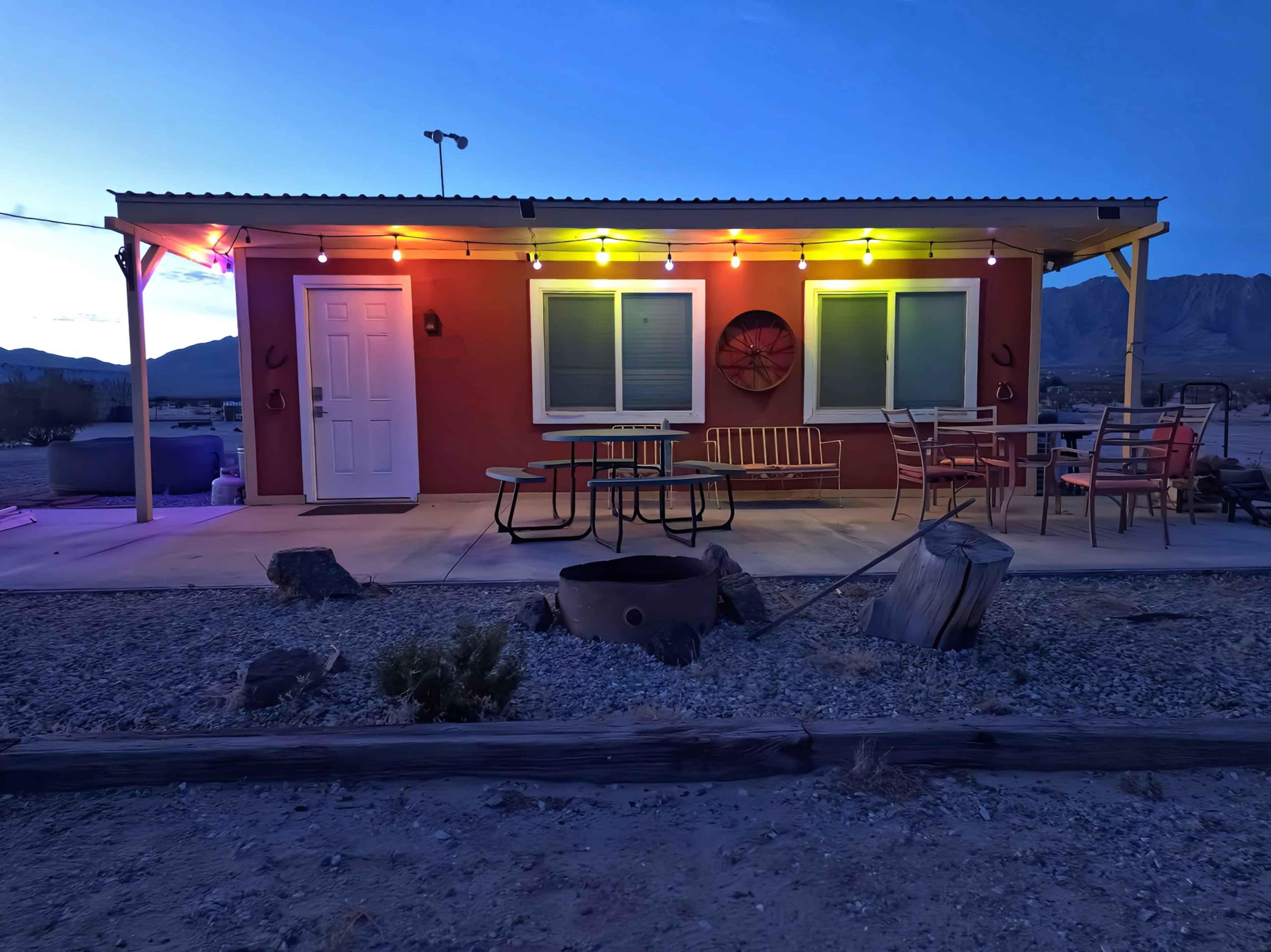 A small, brightly colored outdoor setting features a red building with string lights, a picnic table, and chairs, set against a mountain backdrop at dusk.