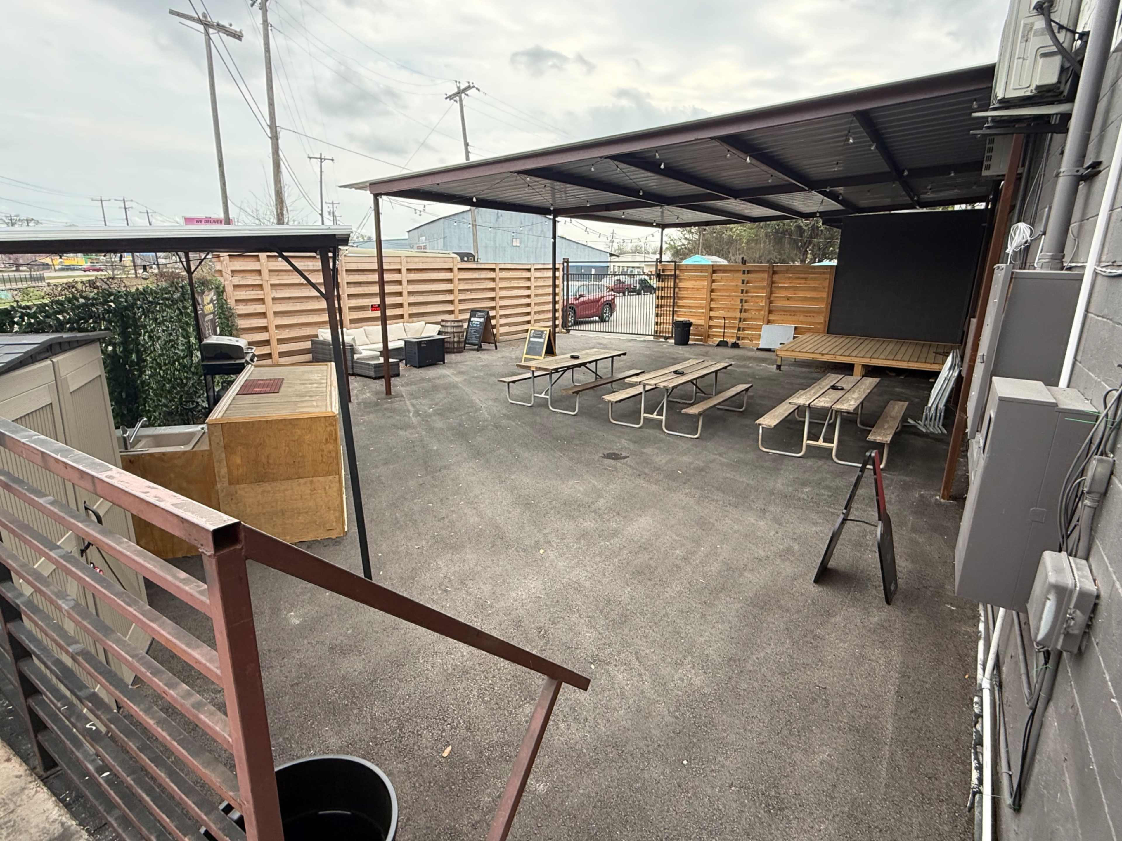 The image shows an outdoor seating area with several wooden picnic tables and benches beneath a metal roof, enclosed by wooden barriers, and featuring a gravel surface.