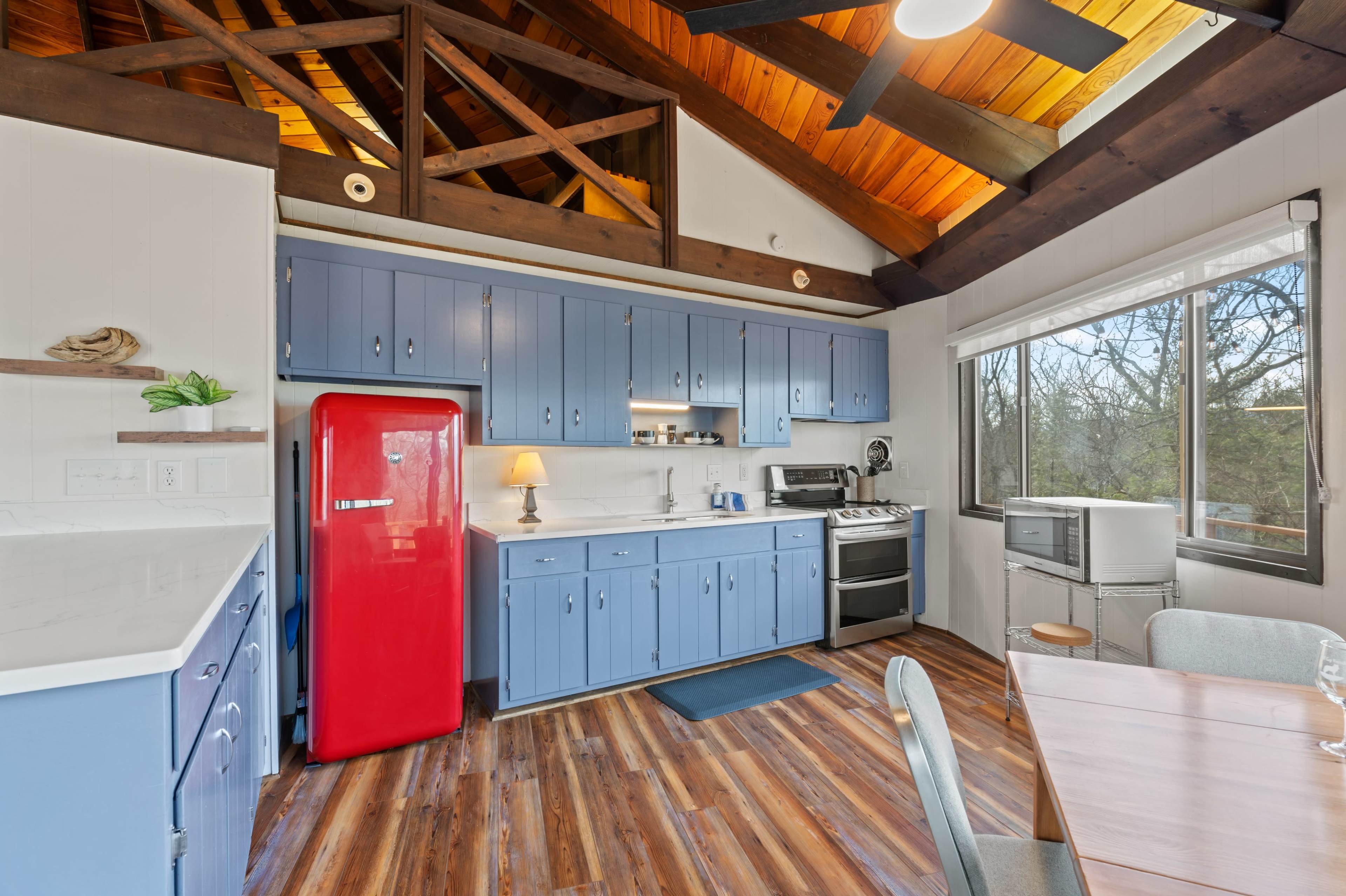The kitchen features blue cabinetry, a bright red refrigerator, and large windows with a view of the outdoors.