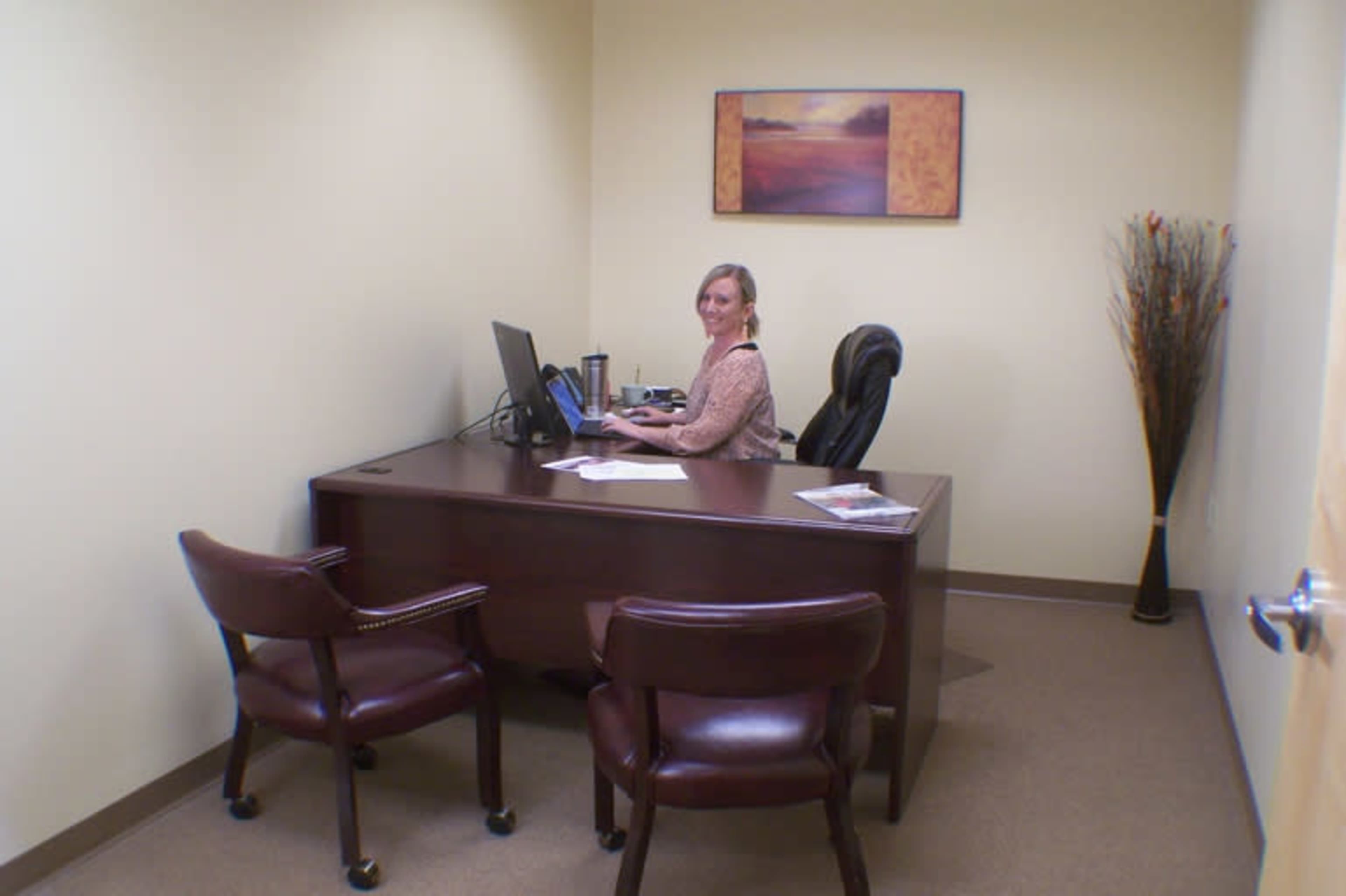 A woman sits at a desk in a small office, working on a computer with two chairs nearby.