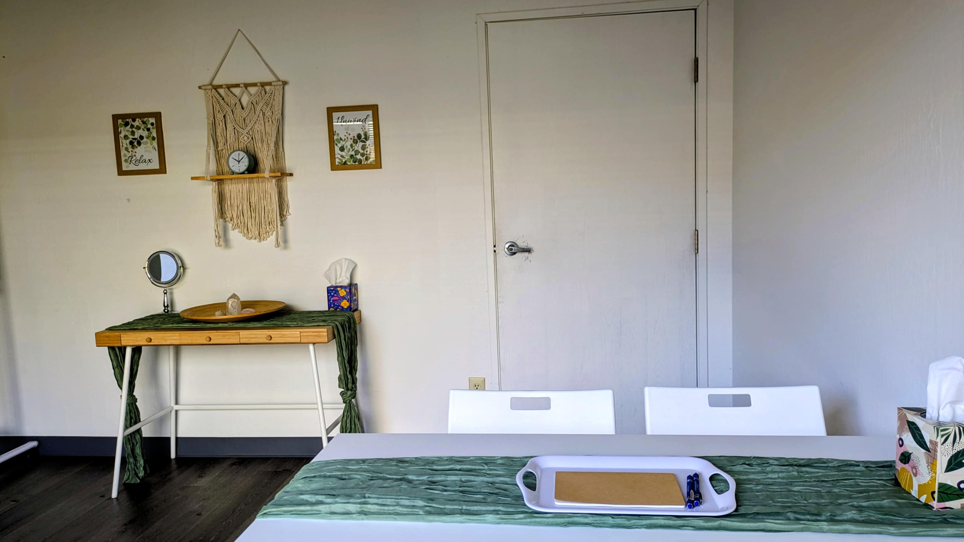 A simple interior scene features a dining table with a green table runner, two white chairs, and a small console table against the wall displaying a mirror, clock, and decorative items.