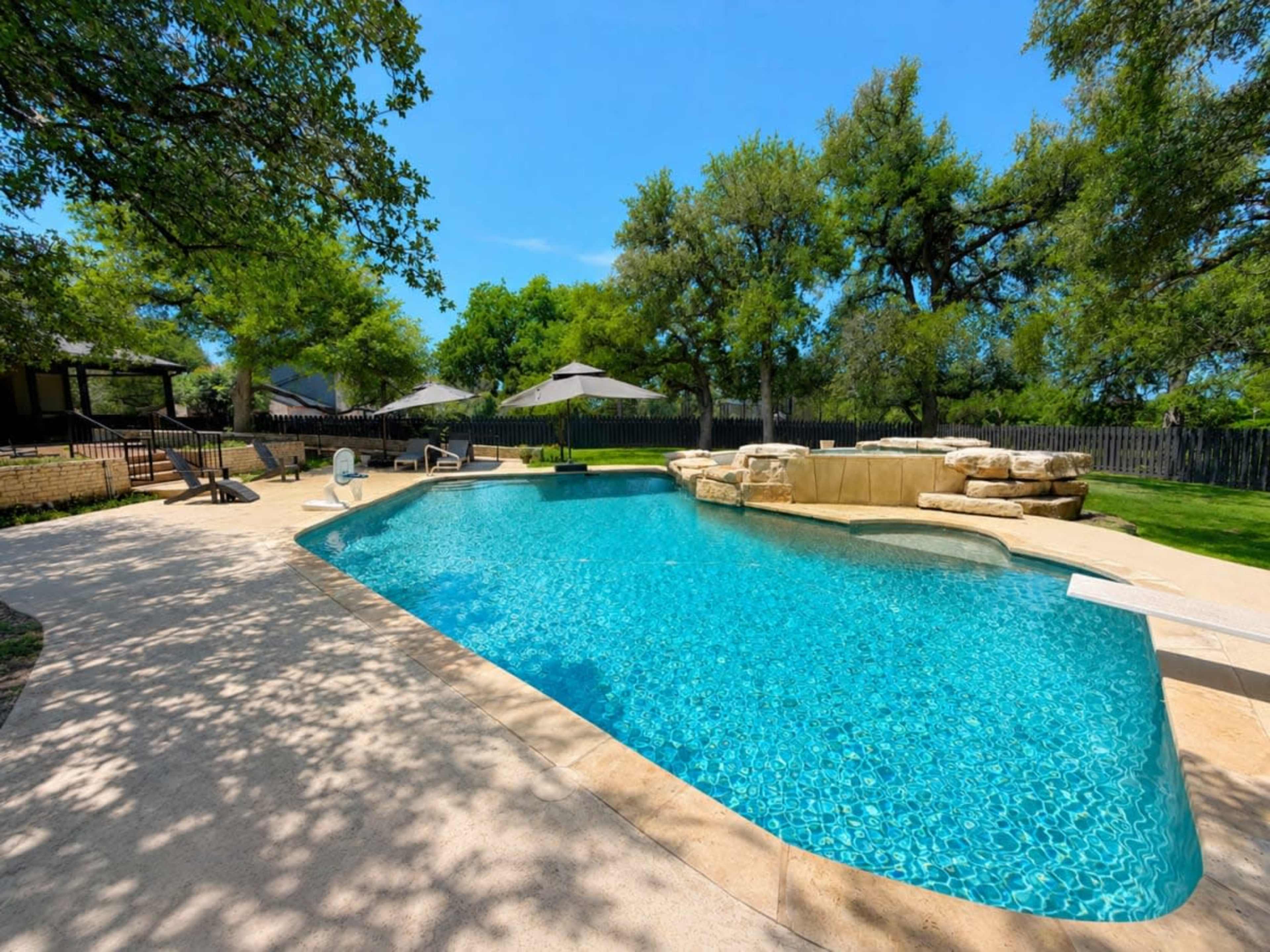 The image shows a landscaped backyard with a swimming pool and surrounding lounge areas under shade from tall trees.