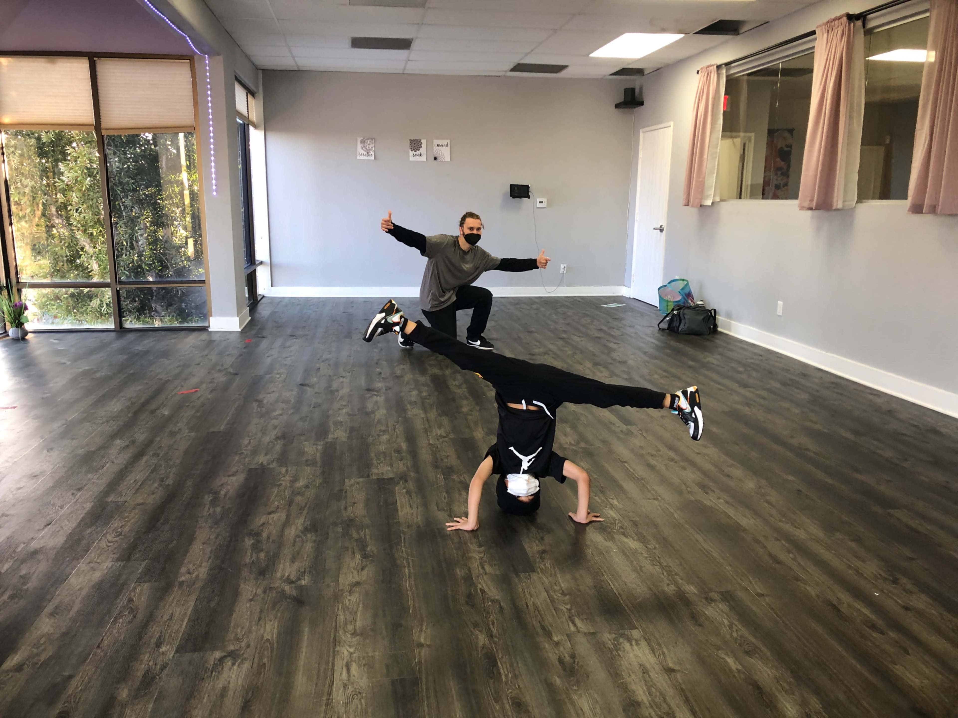 Two individuals are performing dance poses in a spacious, brightly lit studio with wooden flooring.