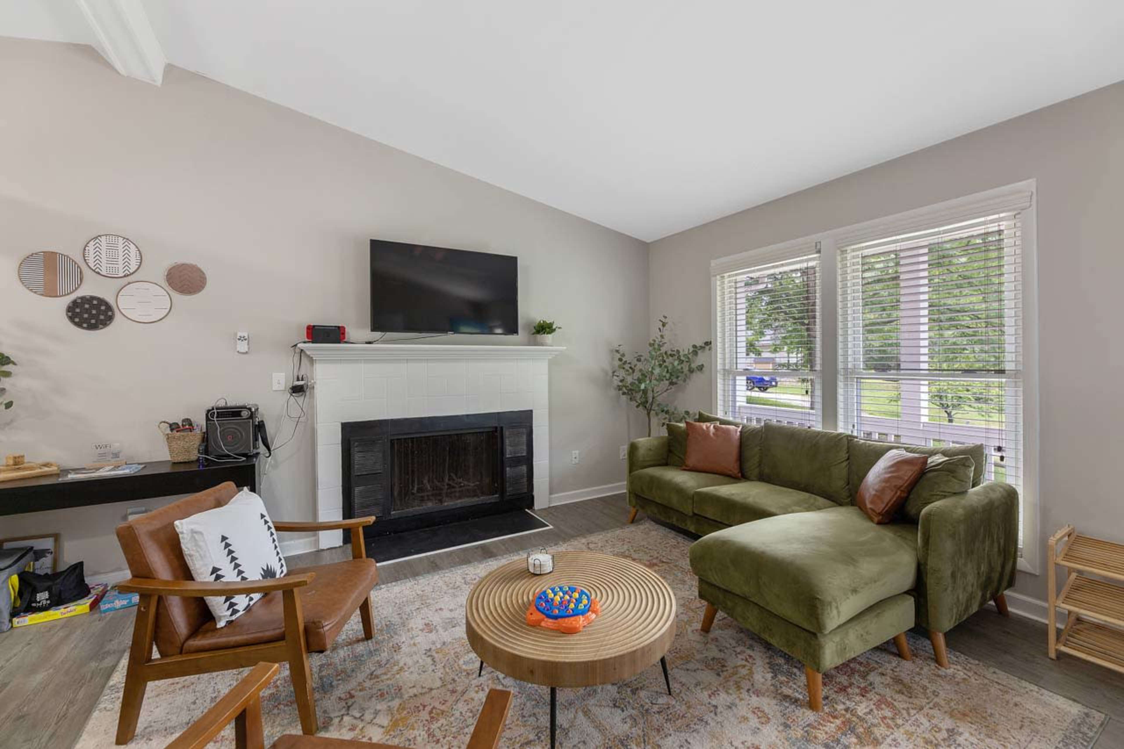 The image shows a living room with a green sofa, wooden armchairs, a round coffee table, a fireplace, and a television mounted above a white mantel.