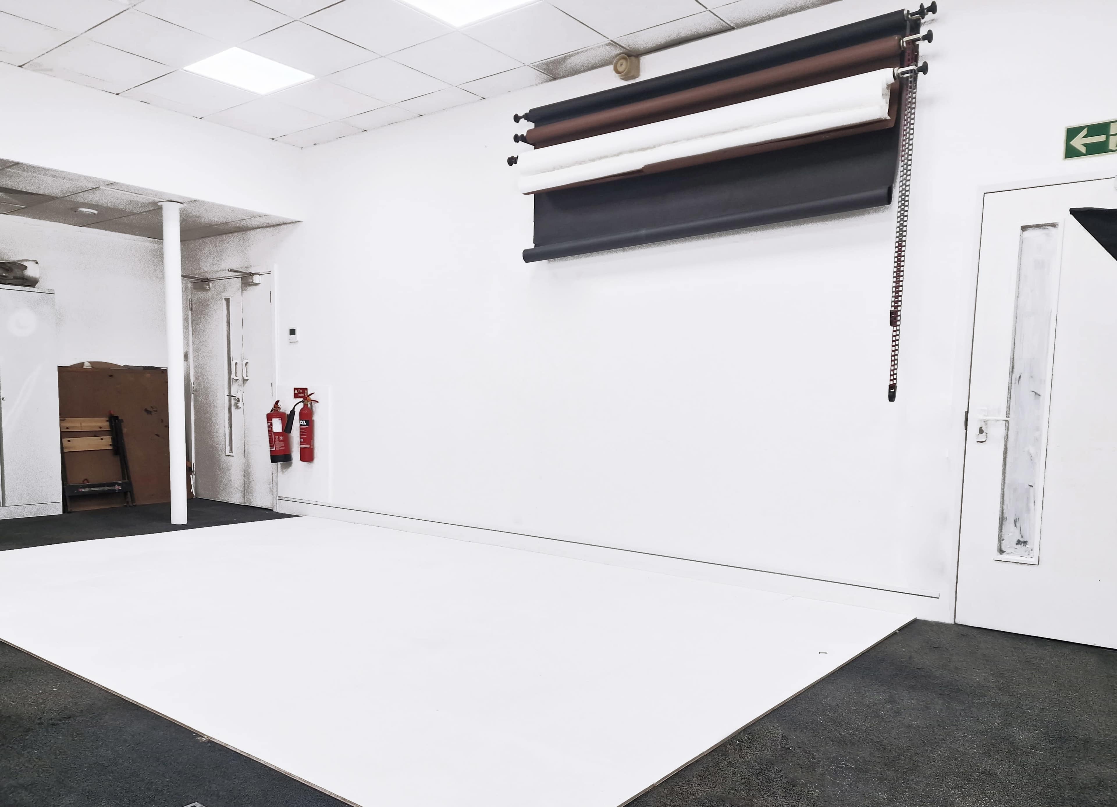 An empty studio space with a white wall and a roll of backdrop paper mounted on the wall, alongside fire extinguishers in the corner.