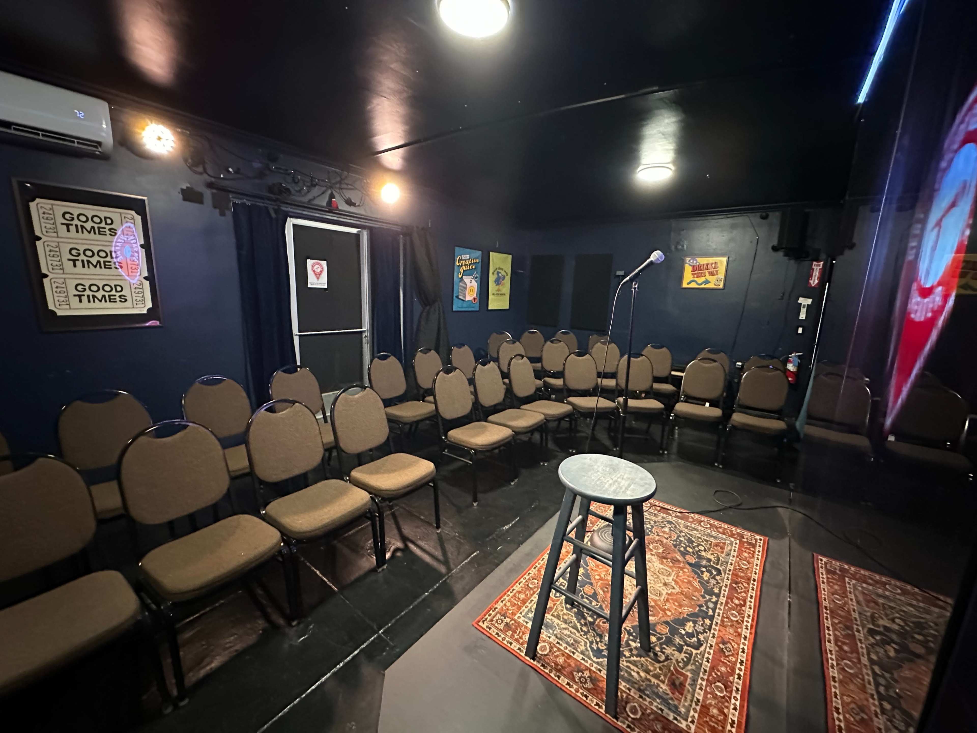 The image shows a comedy club stage with a single microphone on a black stool in front of a row of empty chairs arranged in a semicircle.