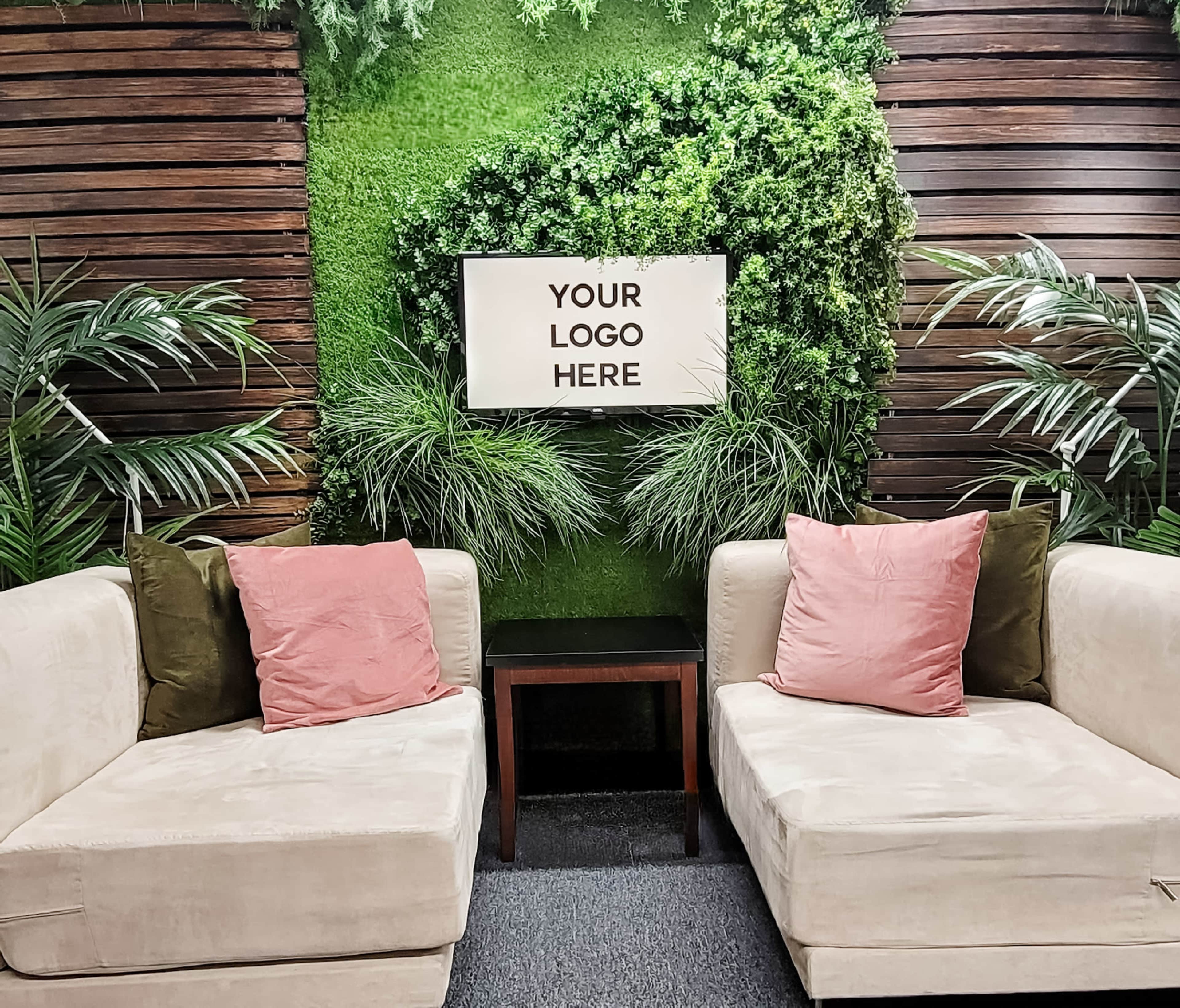 A modern seating area features two light-colored sofas with cushions, a small table, and a green wall adorned with plants and a blank sign reading "YOUR LOGO HERE."
