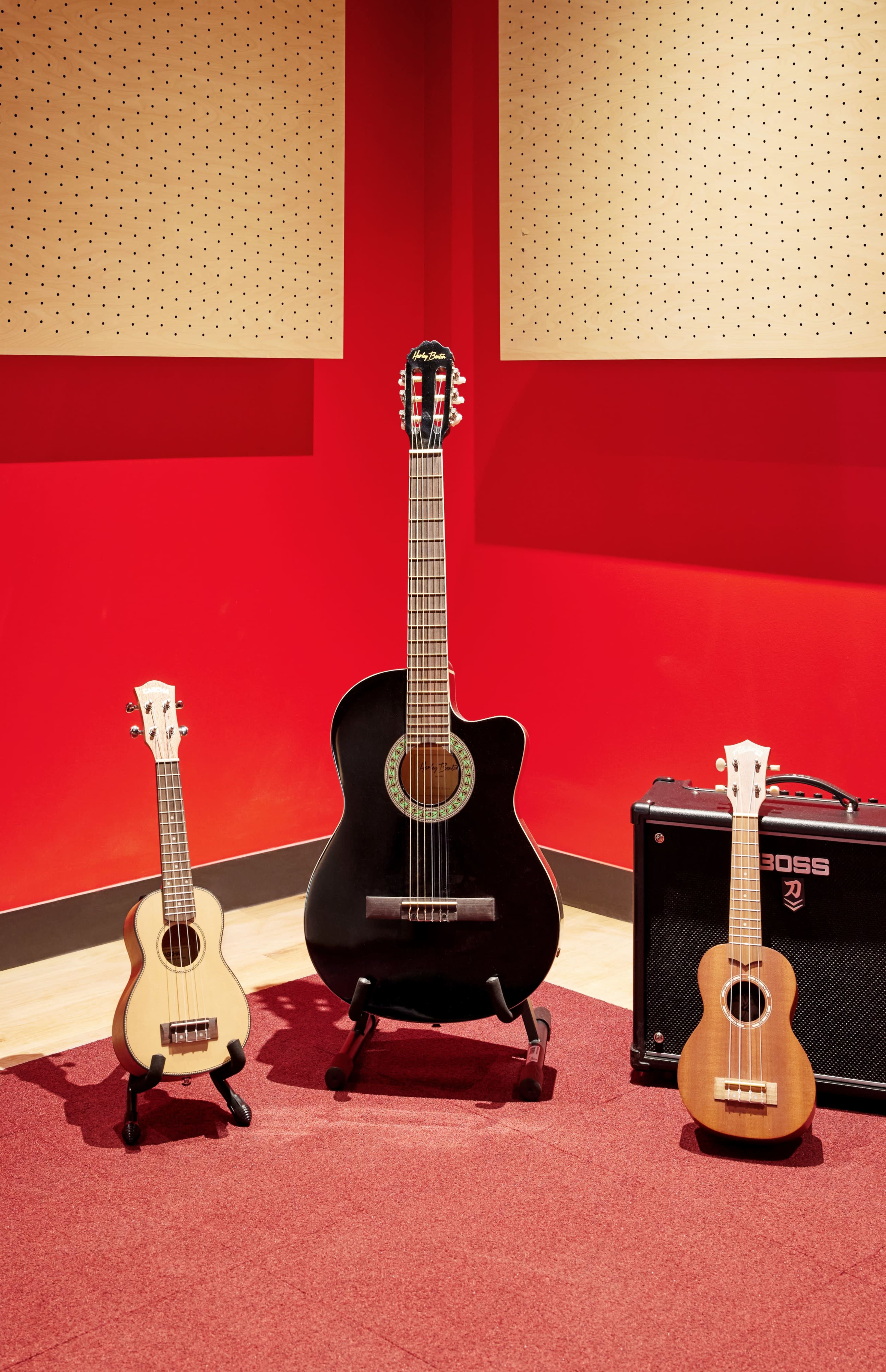 A black acoustic guitar, a small wooden guitar, and a ukulele are arranged in a corner of a room with red walls and a carpet.