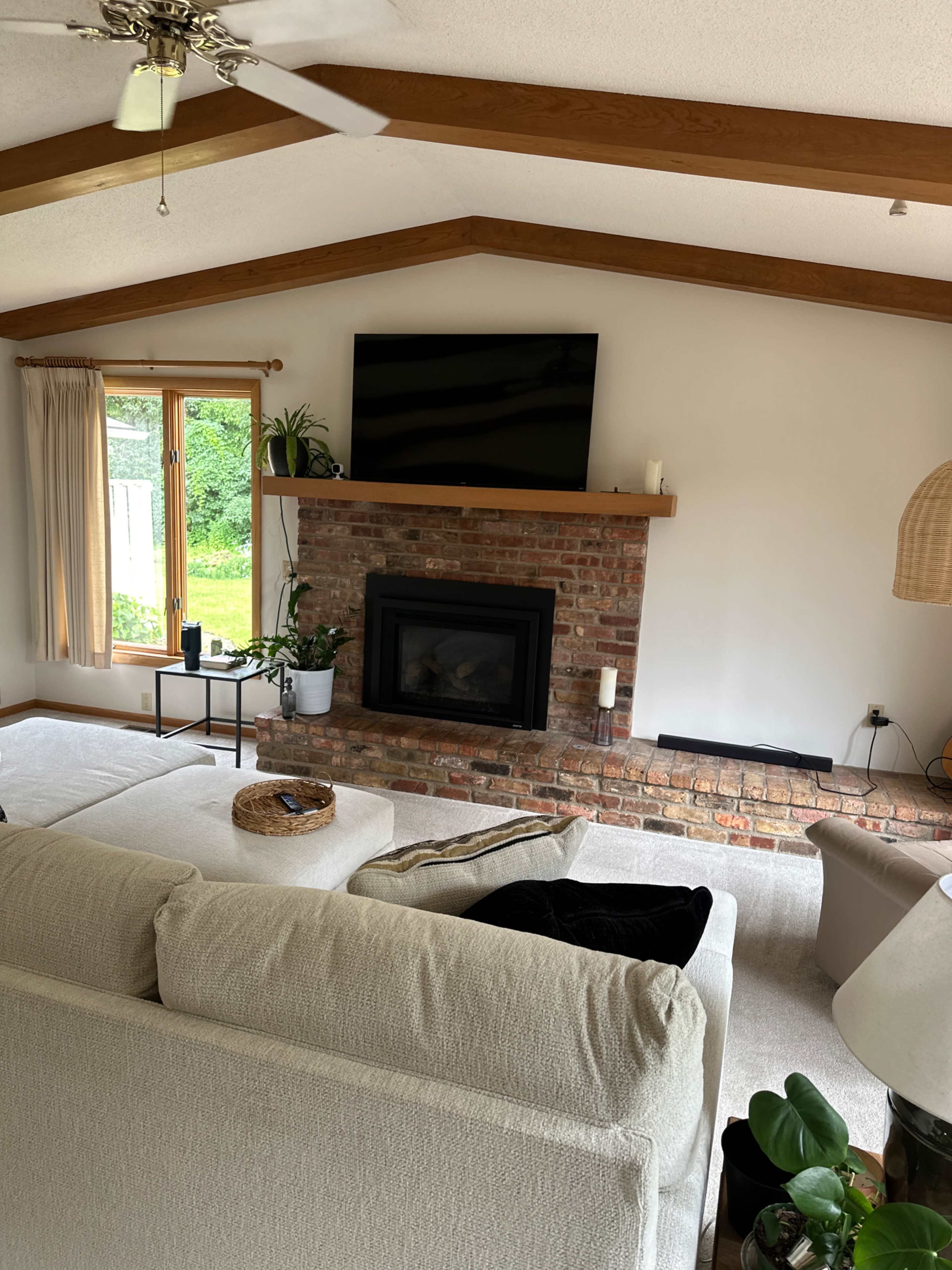 The living room features a brick fireplace with a mounted television above it, a large sectional sofa, and large windows that overlook a green outdoor view.